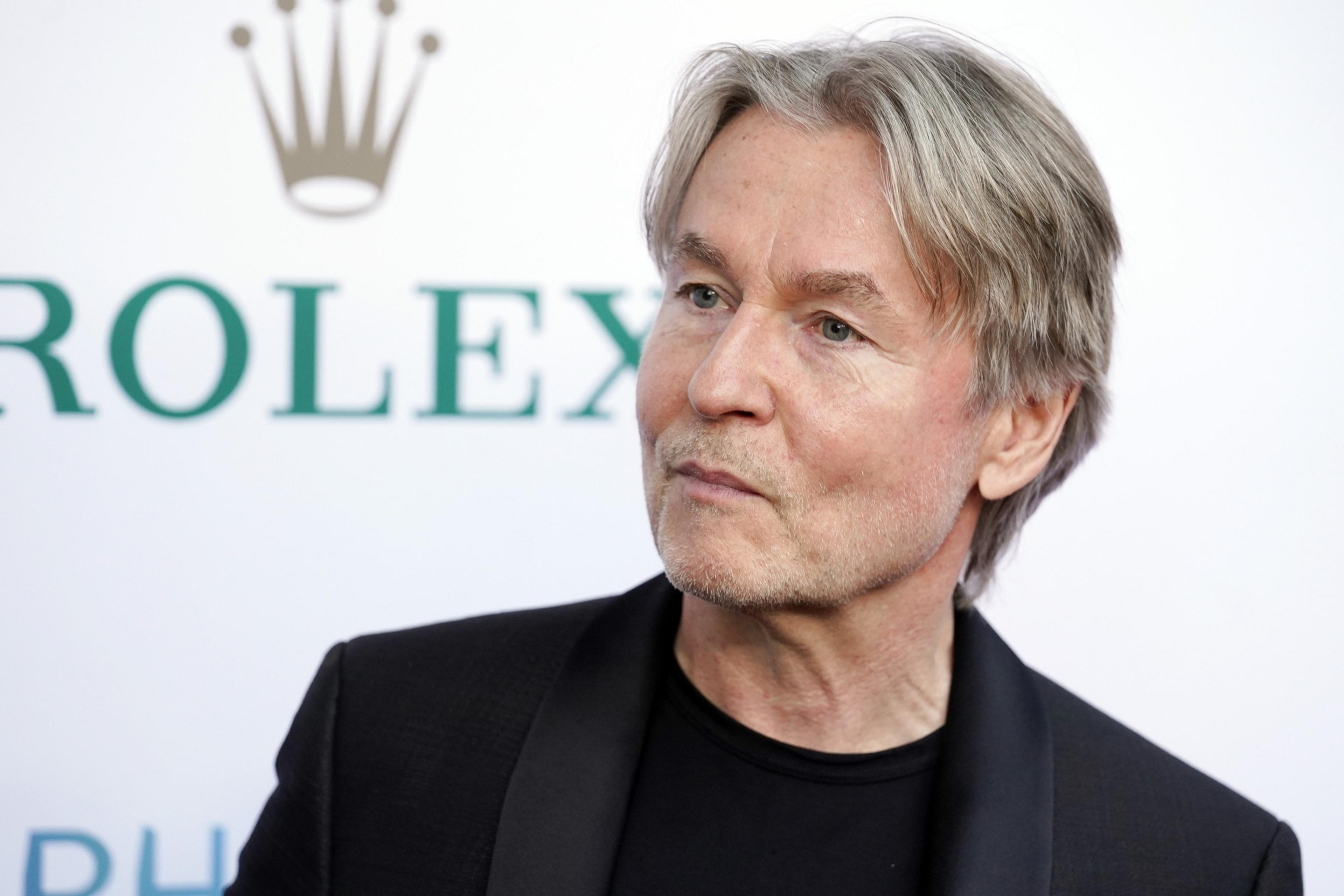 A man with grey hair and a black outfit in front of a blurred Rolex logo.