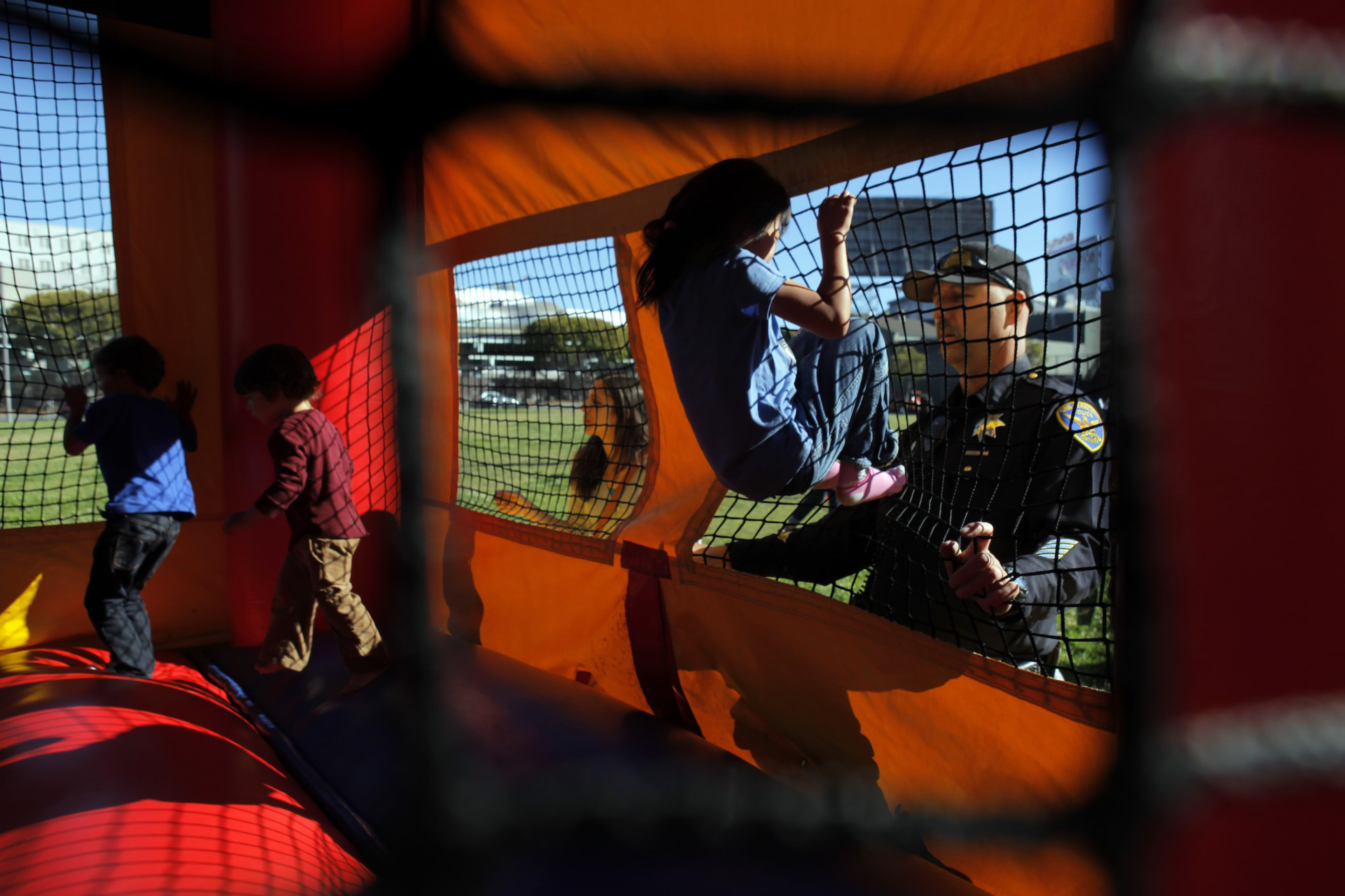 Children play inside an orange inflatable bounce house while a police officer outside helps a girl climb through a netted opening.