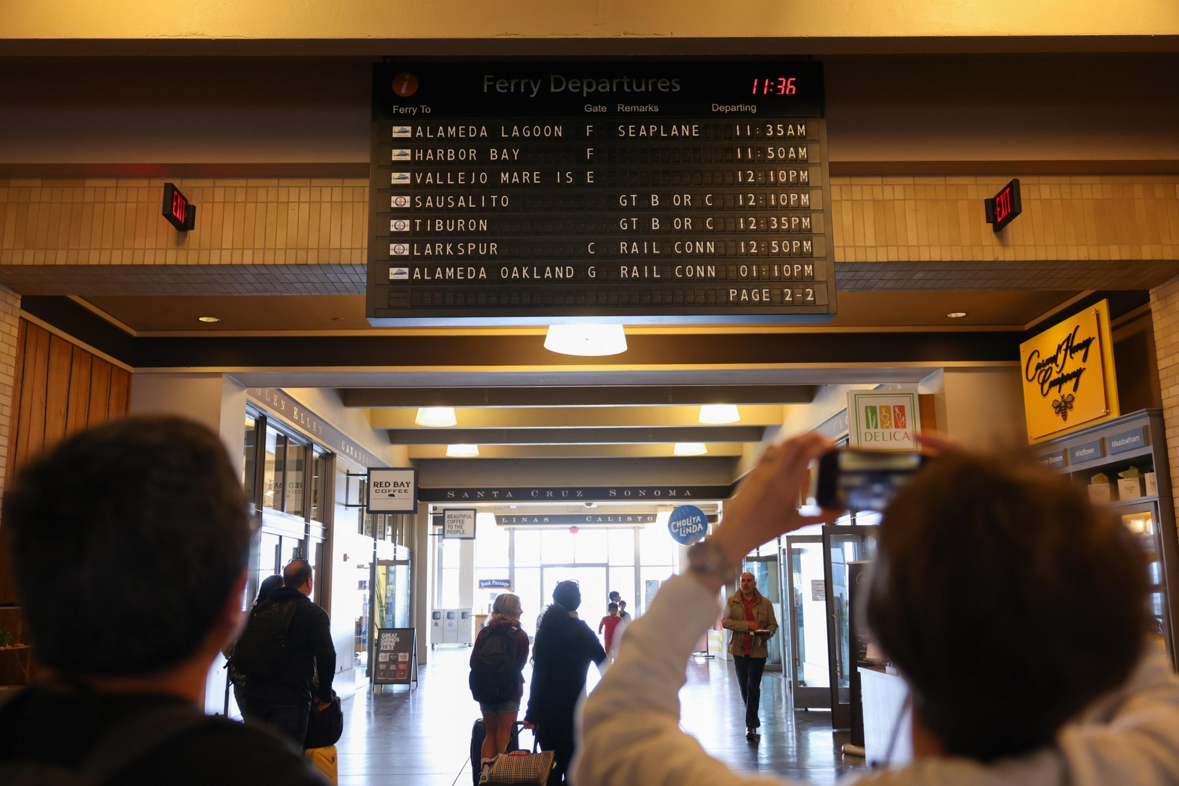 A ferry departure board displays times and gates for several destinations while people look on and one person takes a photo of the board.