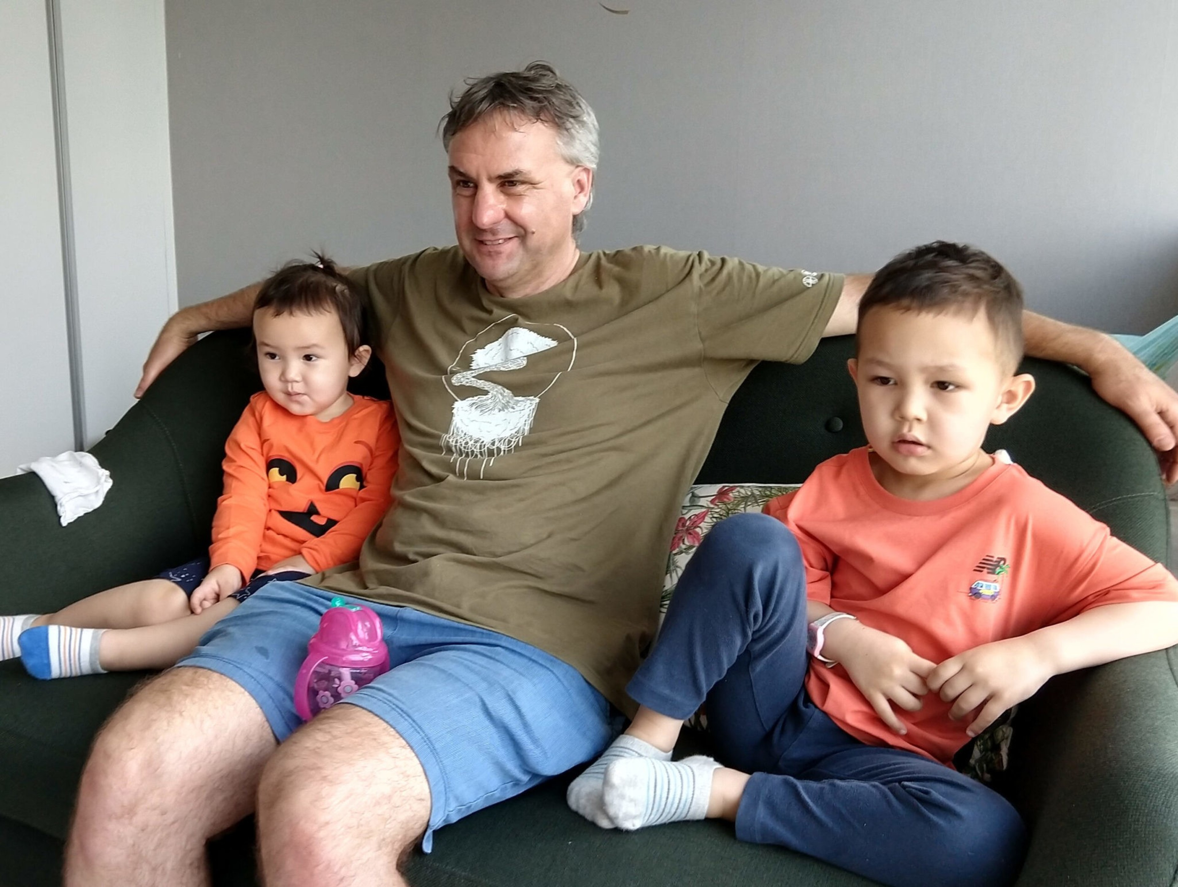 A man in a green shirt and blue shorts sits on a couch with two children, one in an orange shirt with a pumpkin face and the other in a coral shirt.