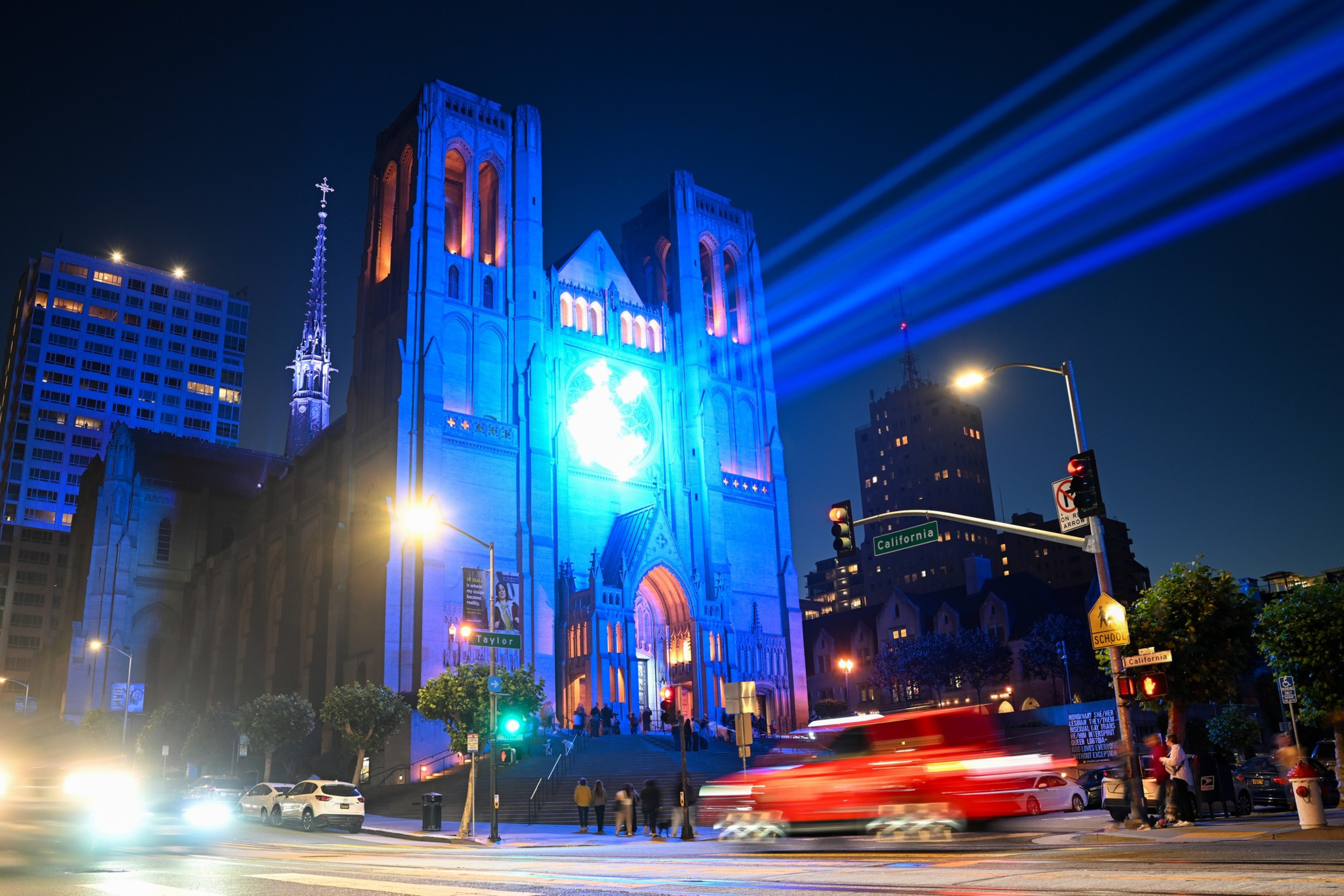 A large gothic cathedral is illuminated in vibrant blue lights at night, with bright beams shooting across the sky and blurred cars passing by on the street.