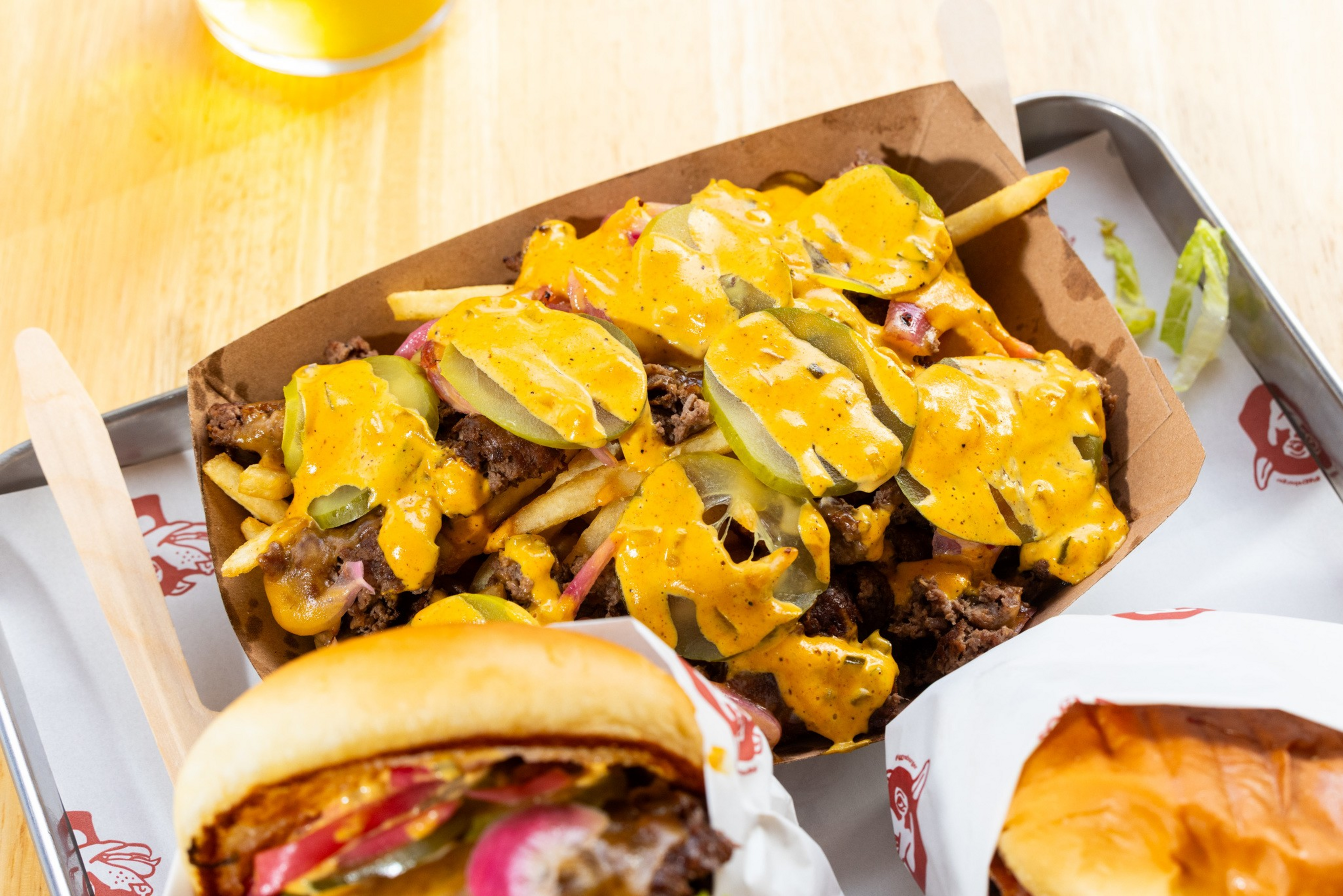 French fries topped with ground beef, pickles, onions, and a generous drizzle of yellow cheese sauce, served on a tray alongside wrapped burgers.