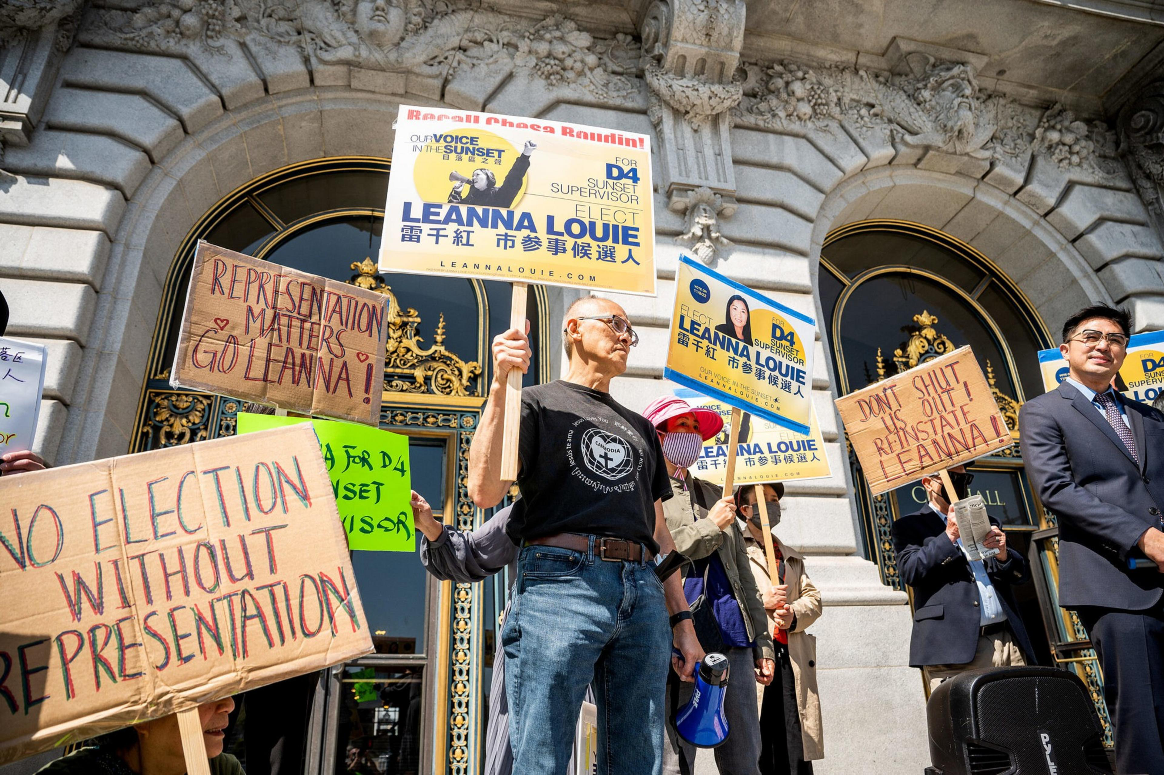 Protesters hold signs supporting Leanna Louie’s election for Sunset District supervisor in front of an ornate building with arches and gold details.