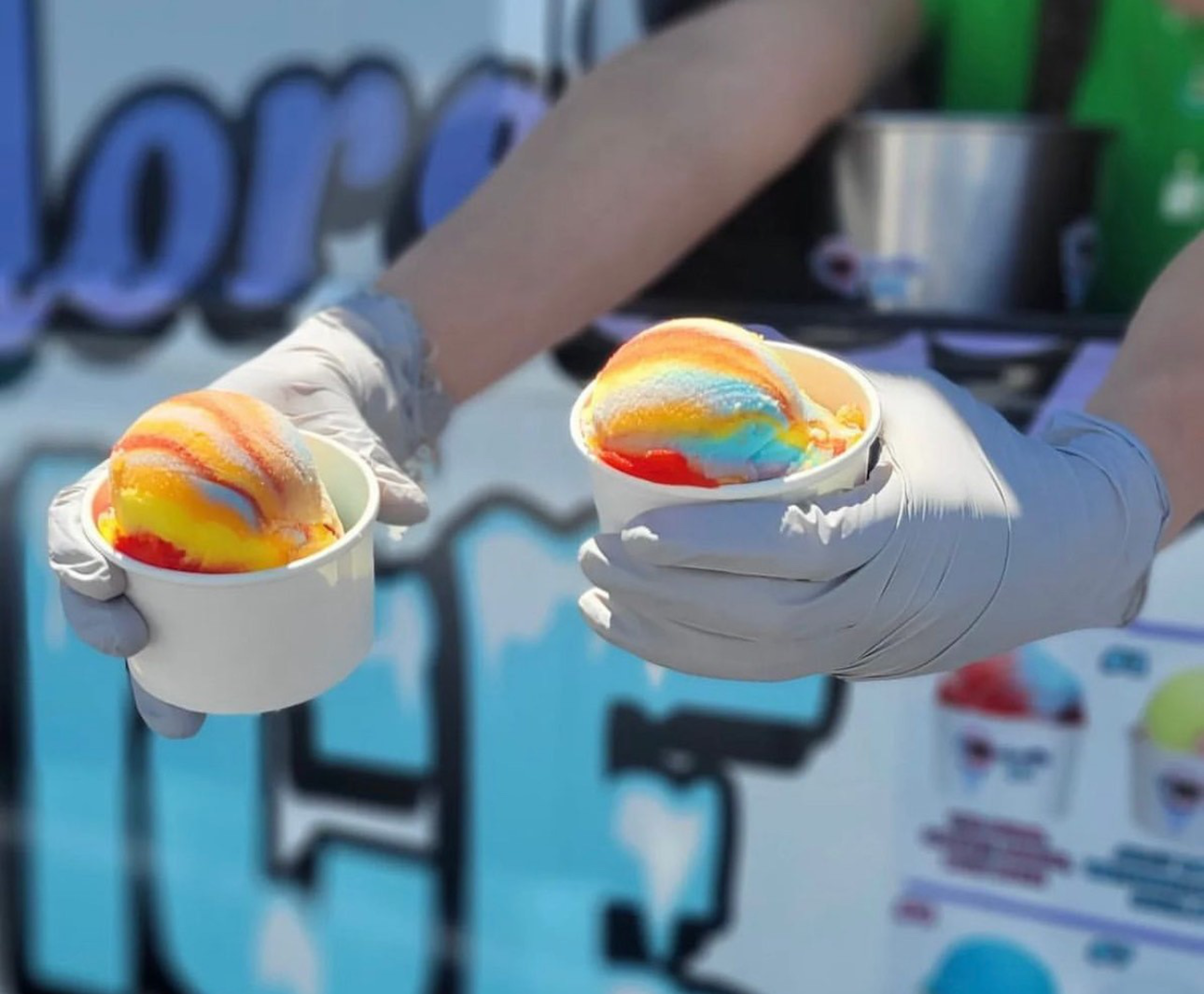 Two hands wearing gray gloves hold small white cups filled with colorful rainbow ice cream scoops.