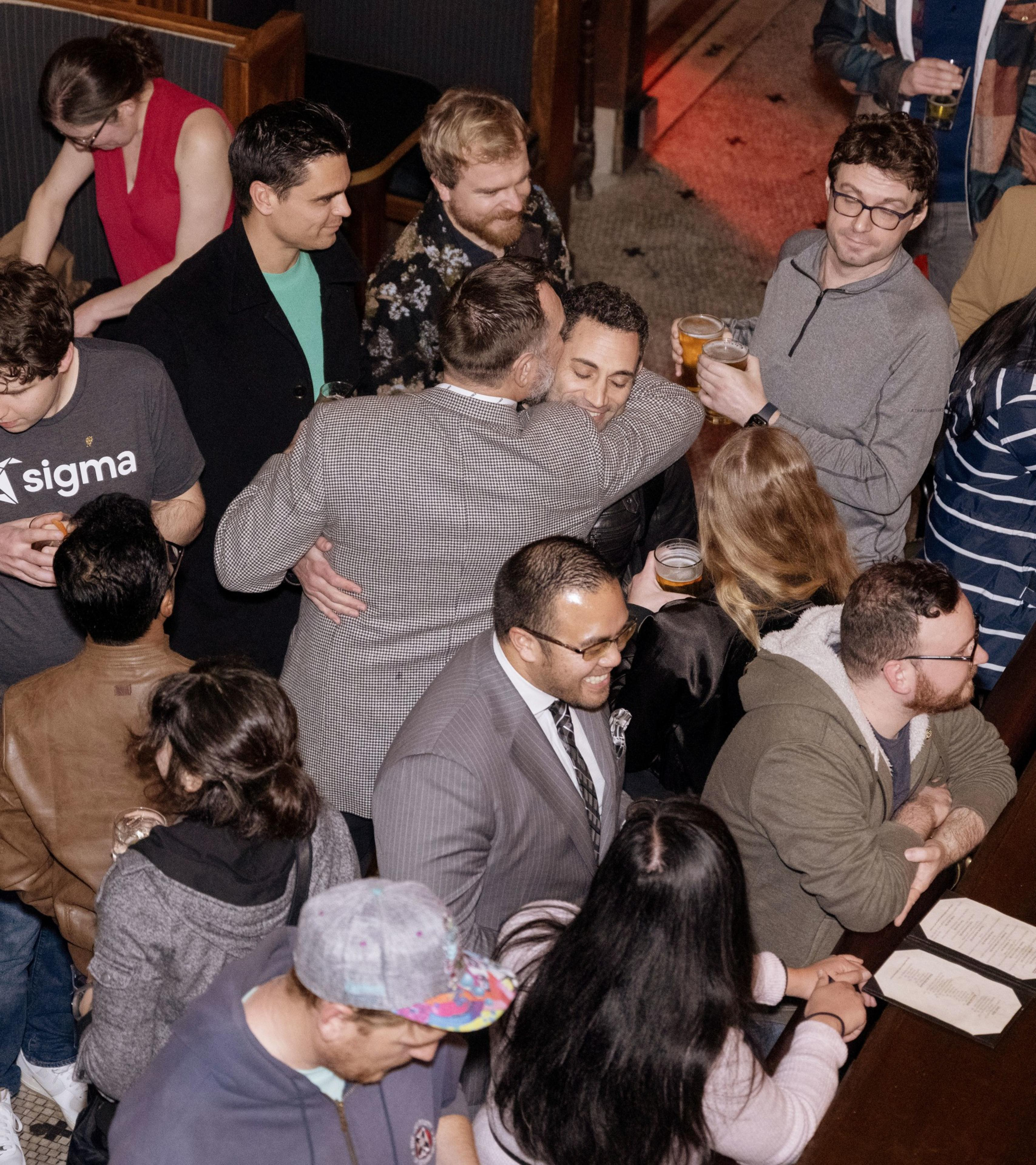 A crowded social gathering with people talking and holding drinks. Some are hugging or laughing, while others are looking at menus or engaged in conversations.