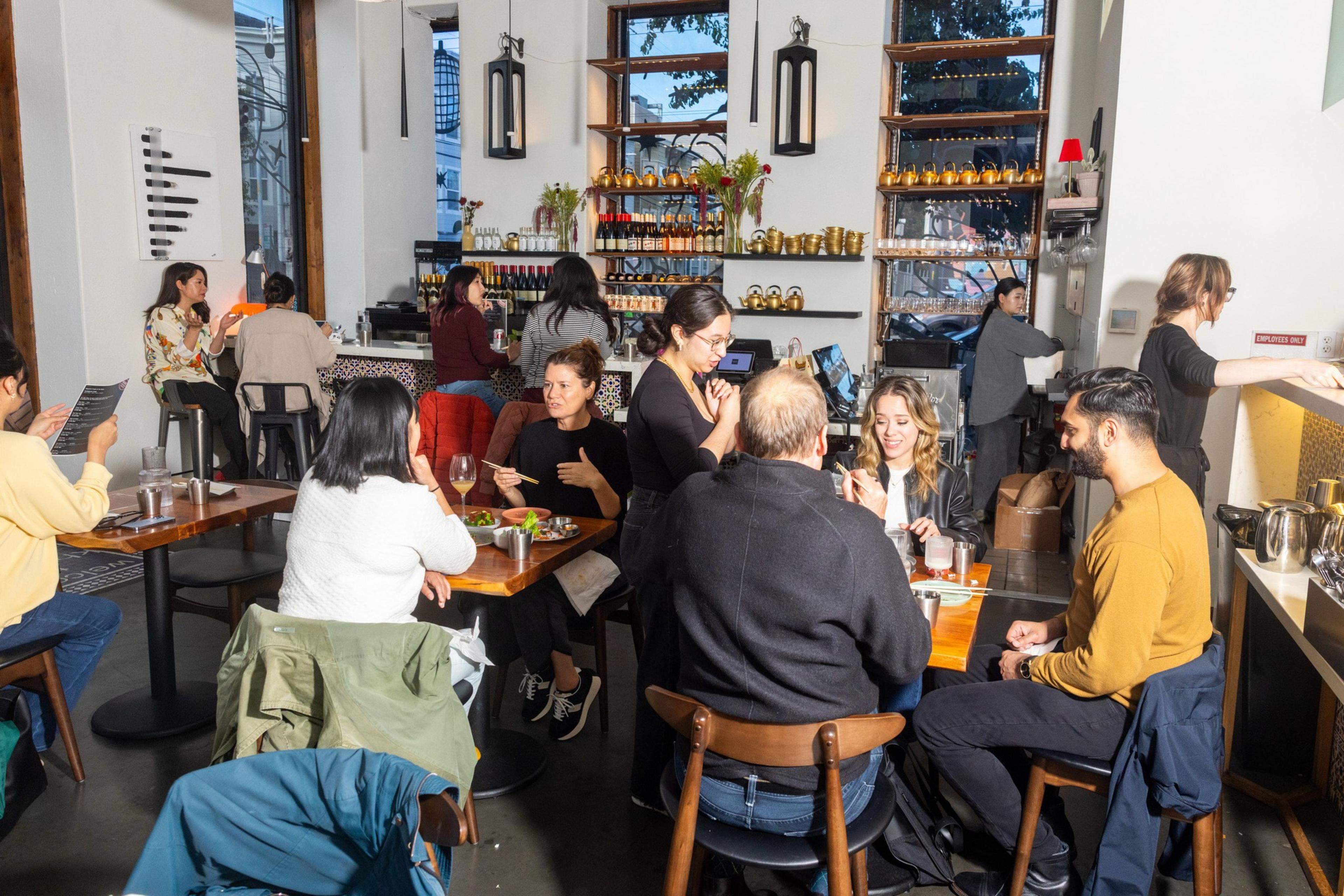 The image shows a lively restaurant with people enjoying meals and chatting. A waiter attends to a table while others sit at the bar. Shelves display bottles and decor.
