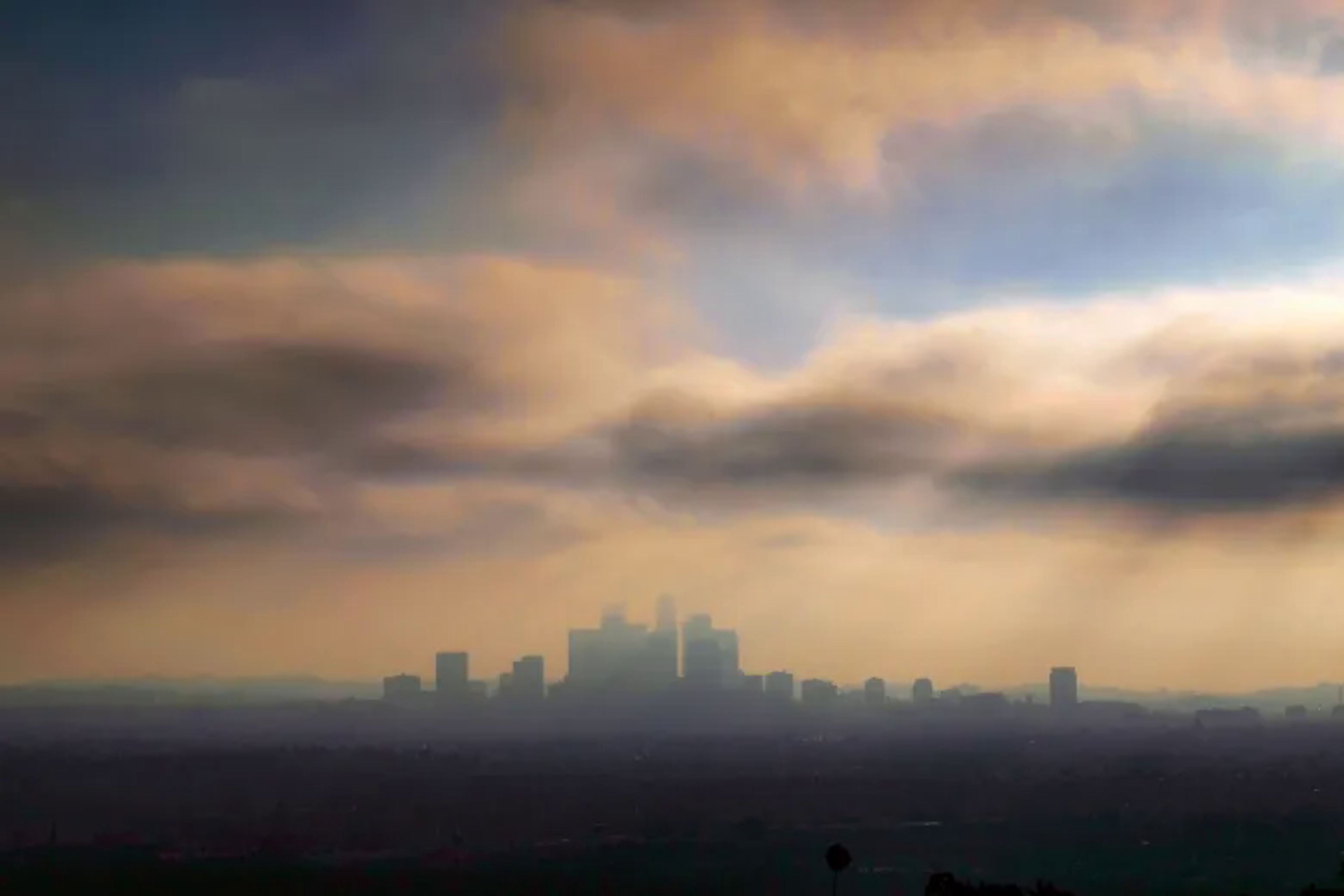 A city skyline is silhouetted against a hazy sky with thick, layered clouds glowing in soft orange and blue hues.