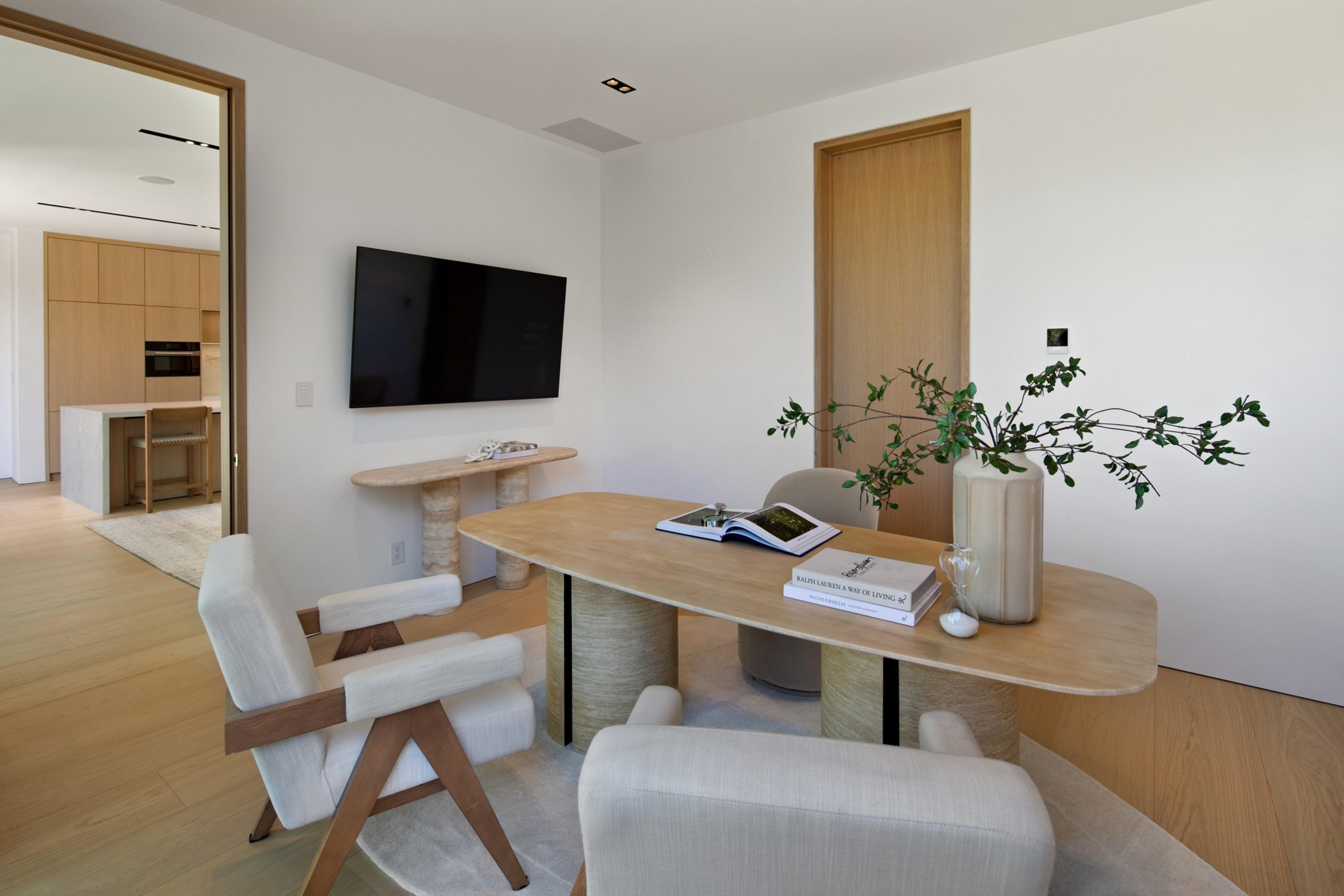 A minimalistic room features a wooden table with books and a vase holding leafy branches, surrounded by neutral upholstered chairs on a round rug.