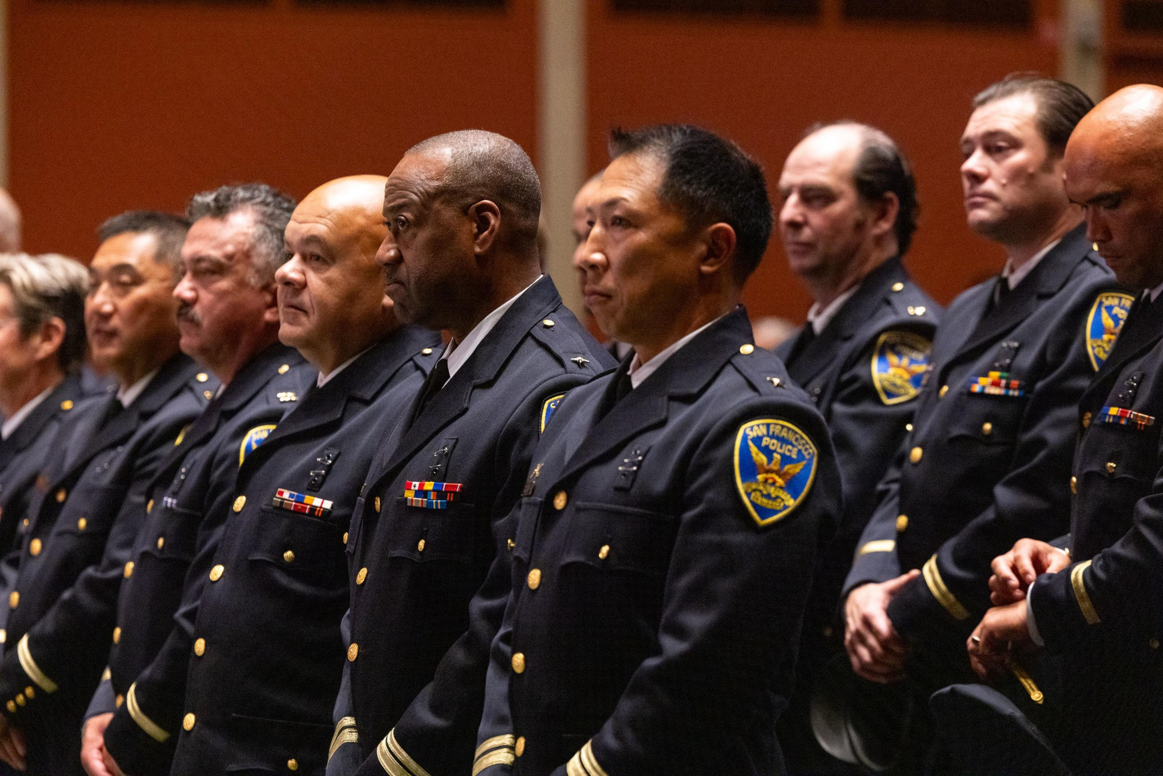 A row of uniformed police officers stands attentively in formation, displaying badges and decorations on their navy blue uniforms, marked with "San Francisco Police."