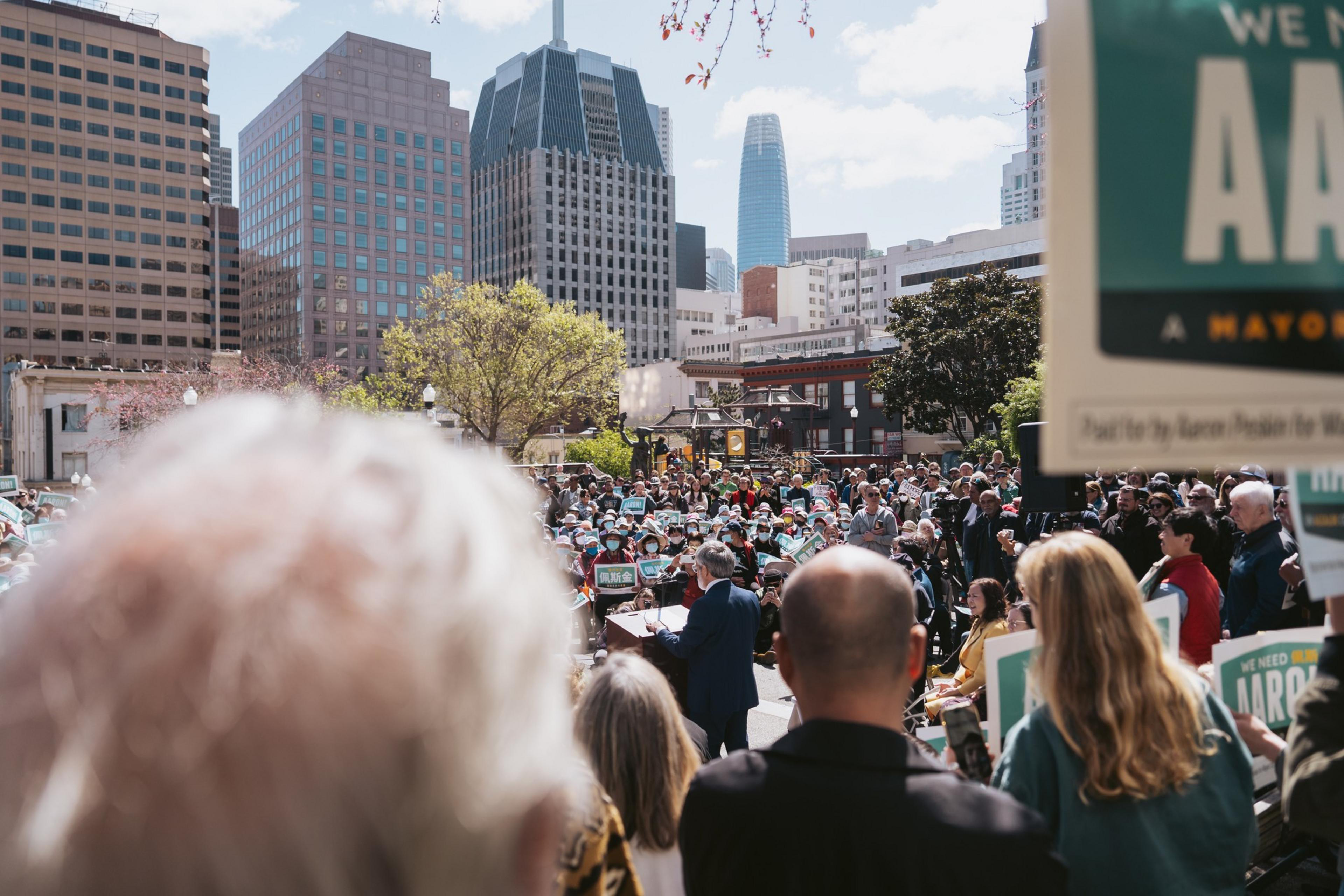 A crowd gathers in a city square, with a speaker addressing them, city buildings in the background.