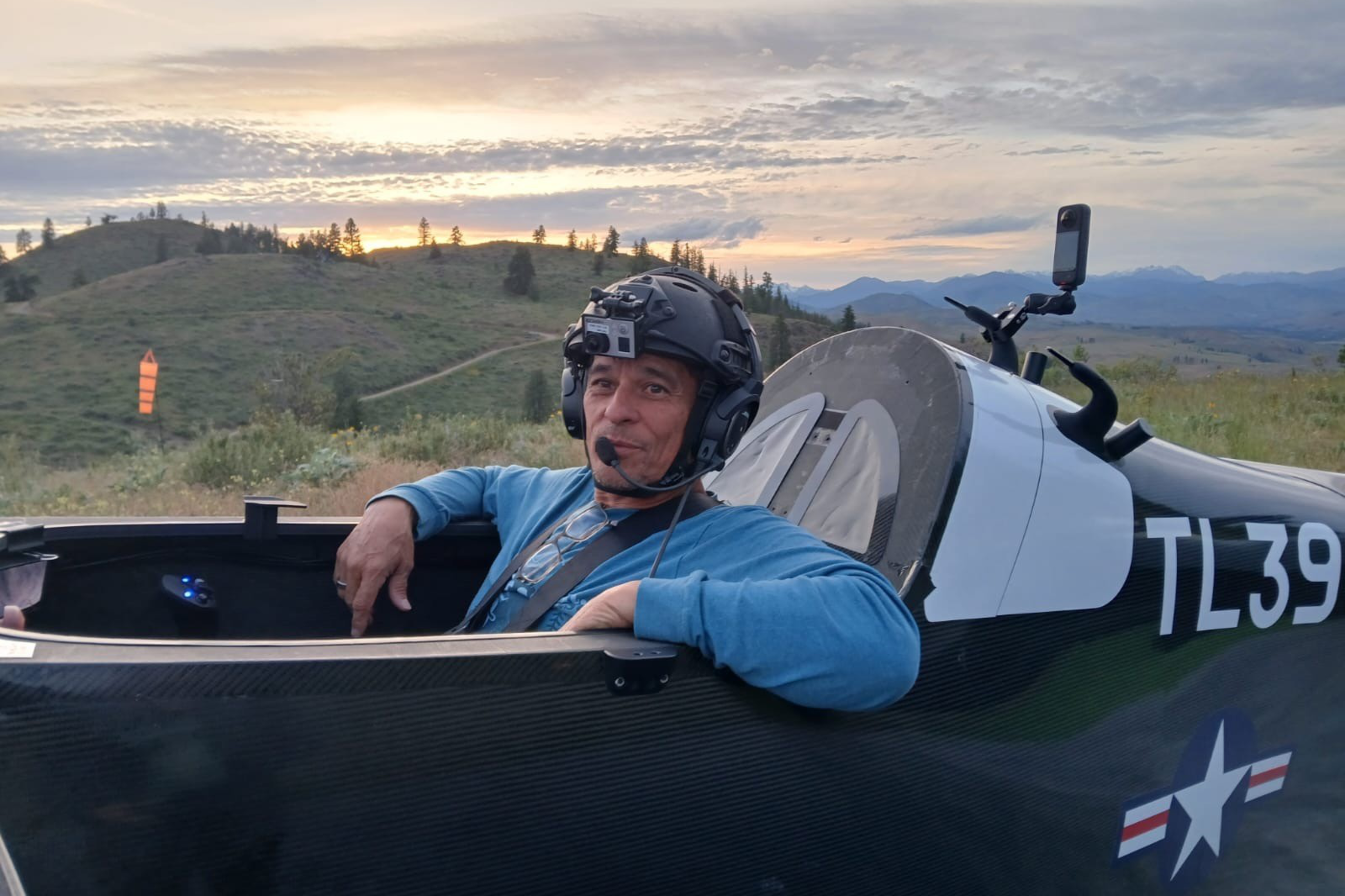 A person wearing a helmet with a mounted camera and headset sits in a black glider cockpit marked with "TL39." The background shows a hilly, scenic sunset.