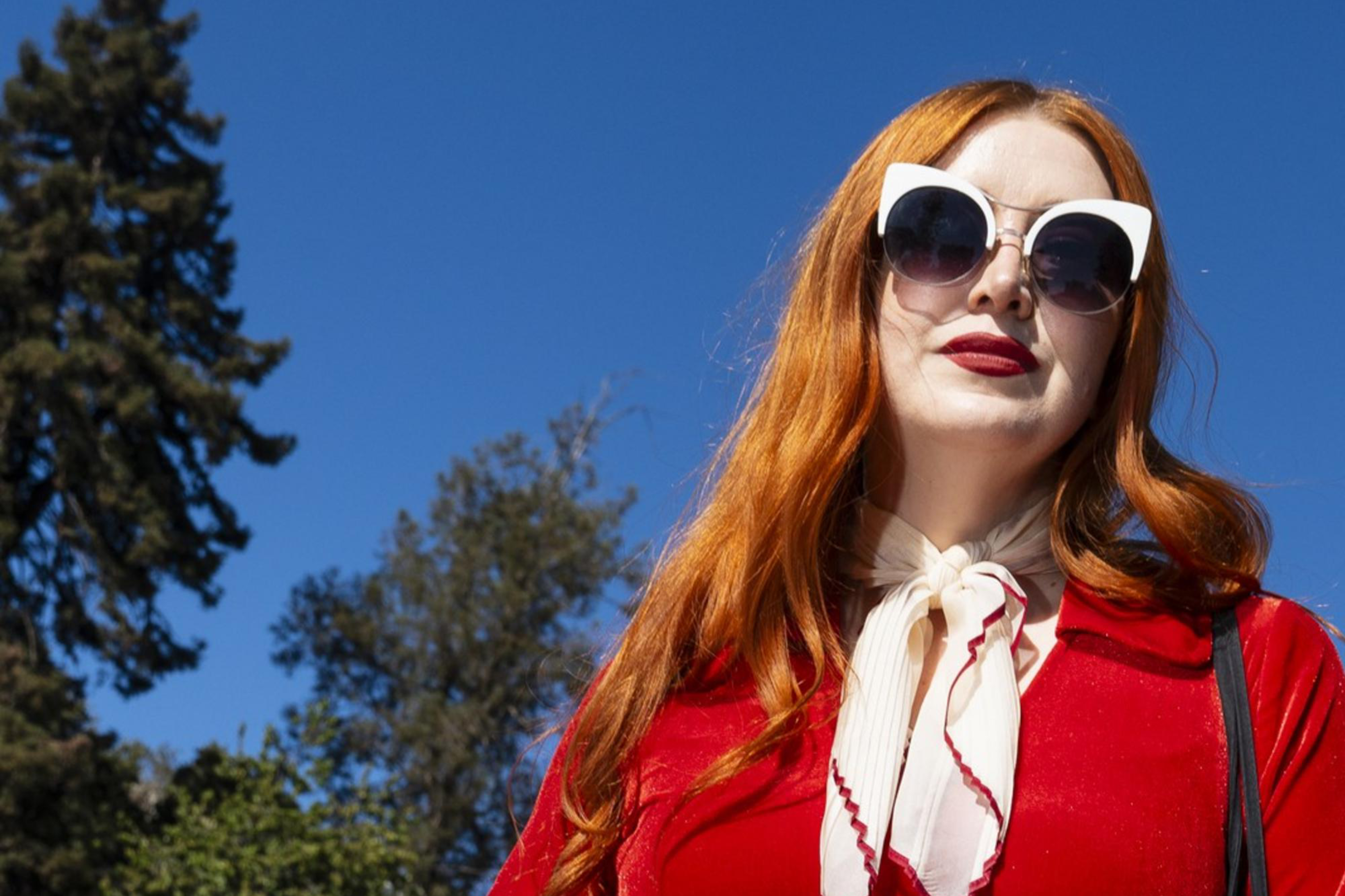 A person with long red hair wears a vibrant red outfit with fringe, a white scarf, heart belt, and sunglasses, standing outdoors with trees and a blue sky.