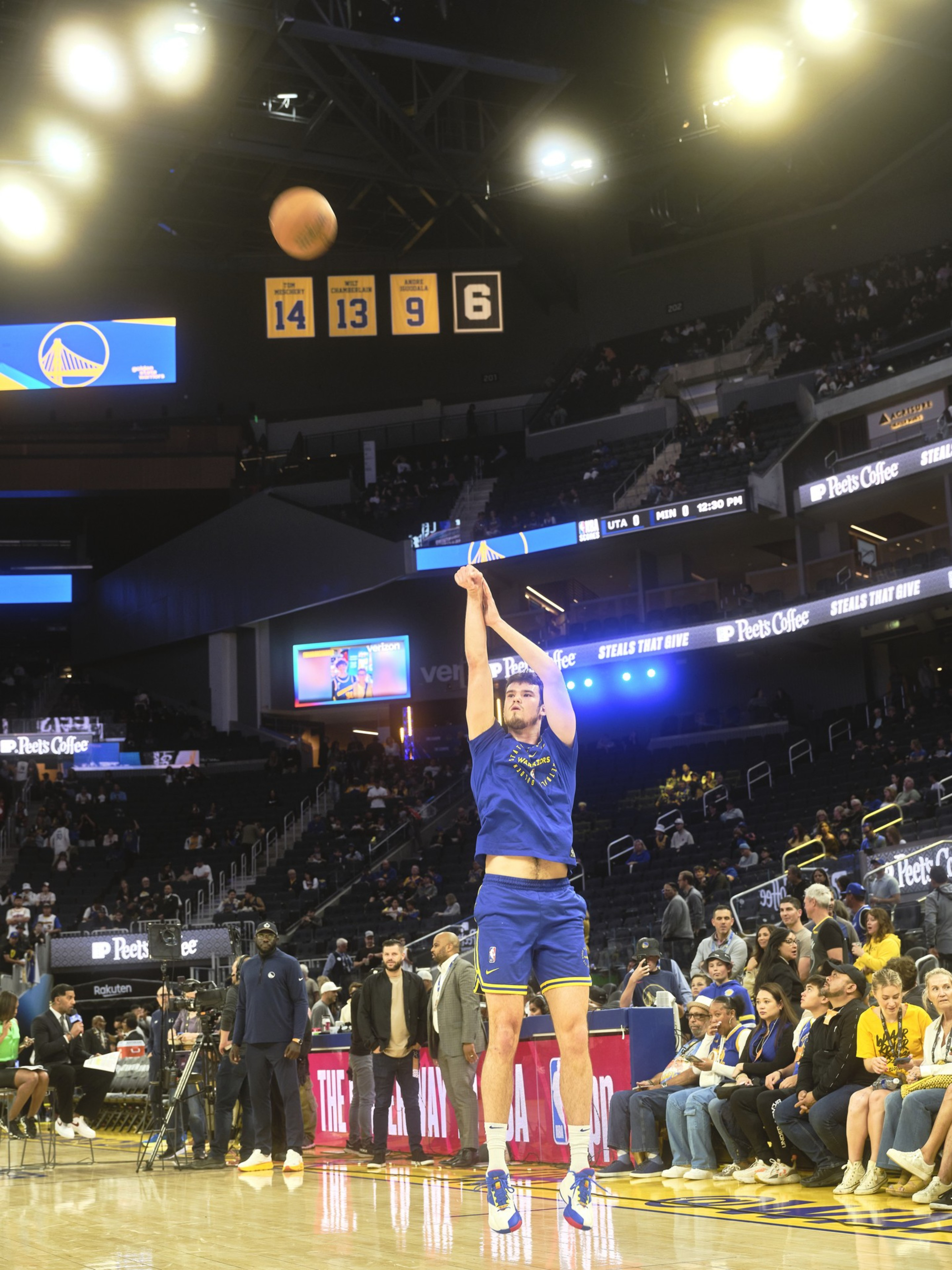 A basketball player in a blue uniform is shooting a ball mid-air on a court. Spectators are seated around, with banners and lights overhead.