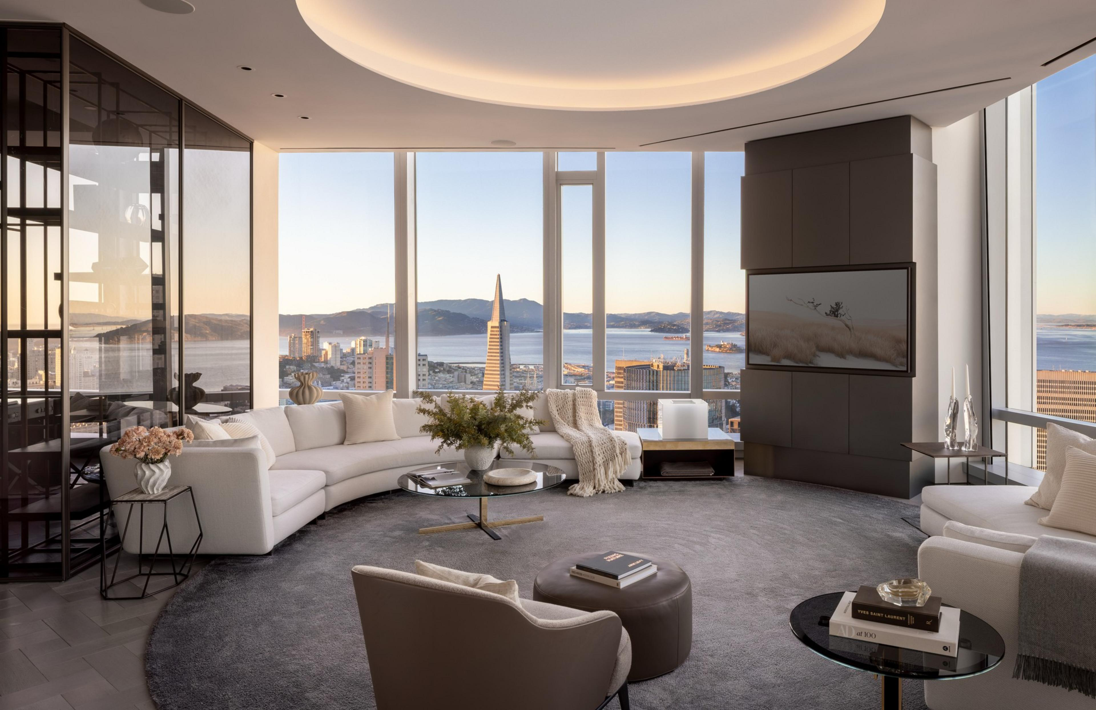 A modern living room with a curved white sofa, large windows showing a cityscape and bay, a round gray rug, and a wall-mounted TV.