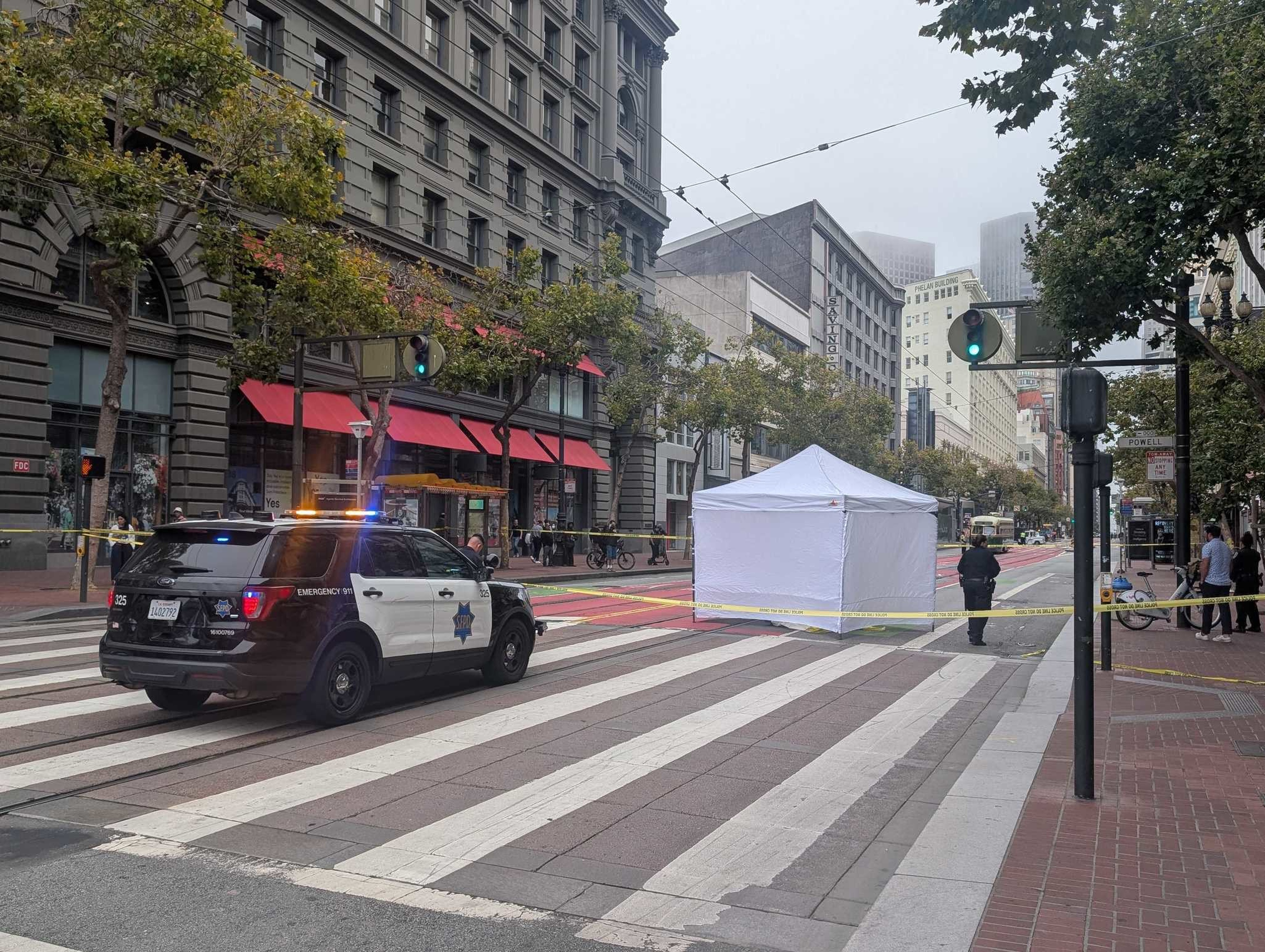 A police car blocks a city street with a white tent and yellow caution tape surrounding an area on a crosswalk, with officers and bystanders nearby.