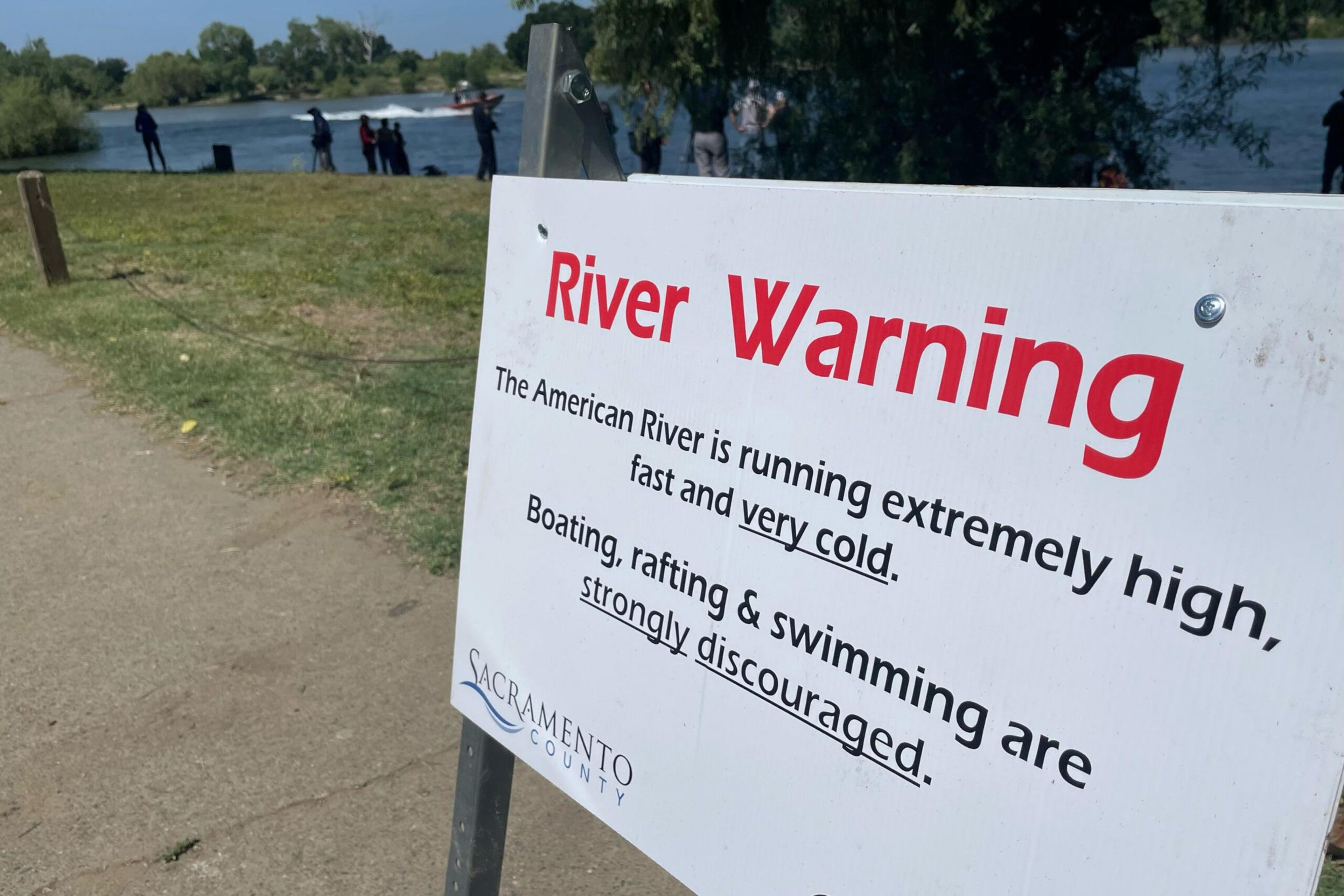 A sign warns that the American River is extremely high, fast, and very cold; boating, rafting, and swimming are strongly discouraged. People stand near the river.
