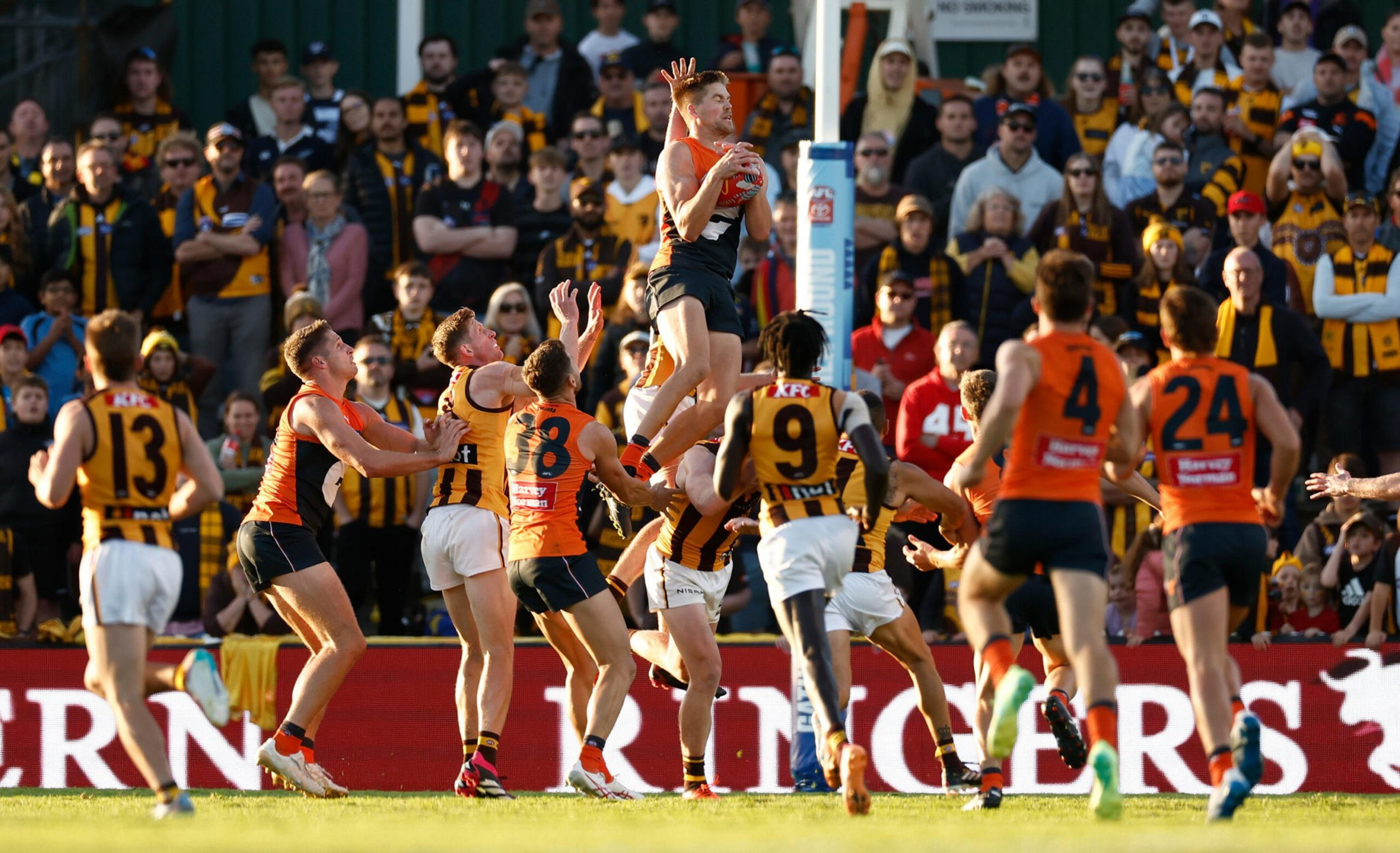 An Australian rules football player in a black and red uniform jumps to catch the ball, surrounded by players in orange and striped yellow-black uniforms.