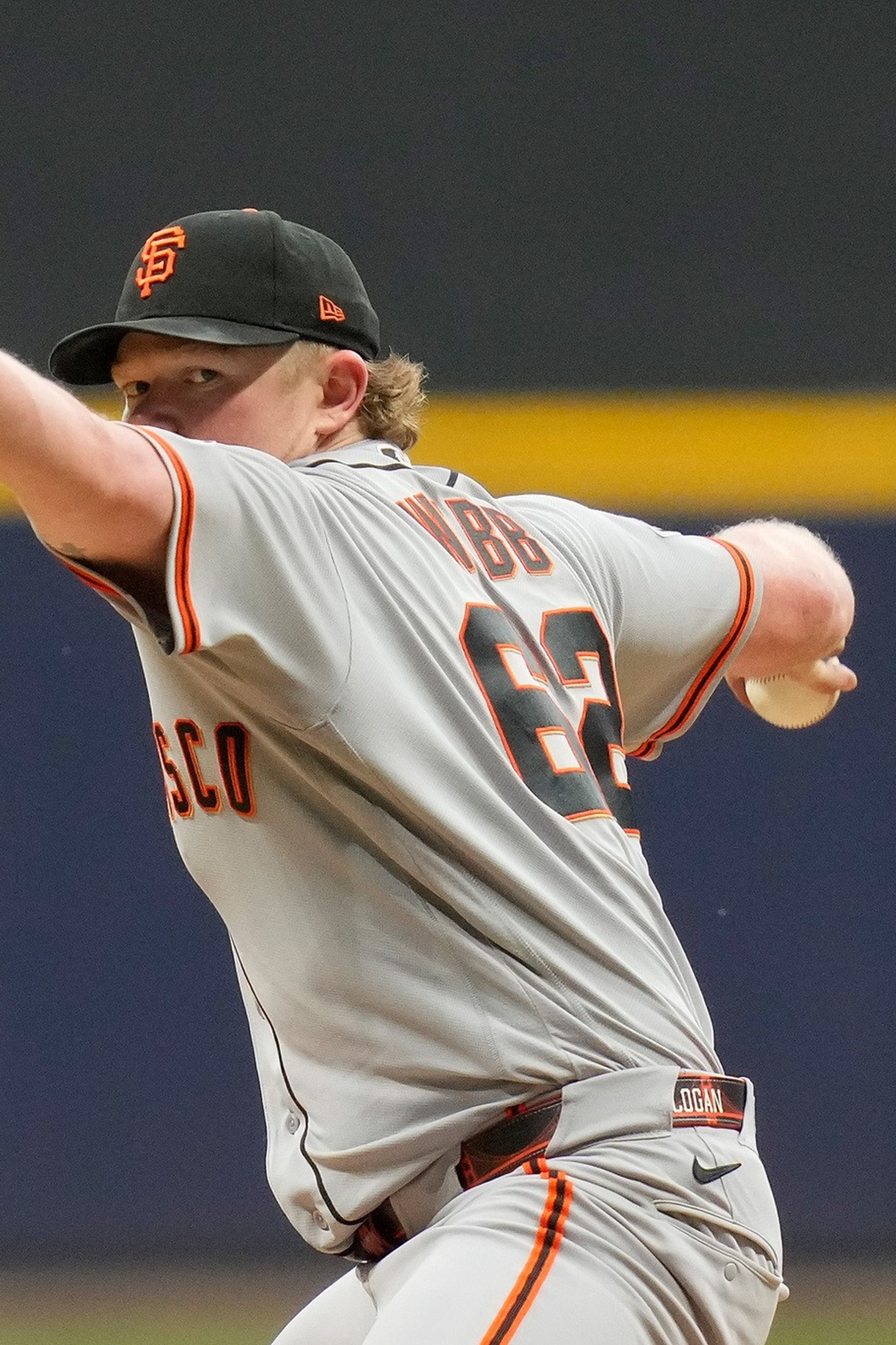 A baseball pitcher in a gray San Francisco Giants uniform is mid-throw, with his glove extended forward and the ball in his other hand.