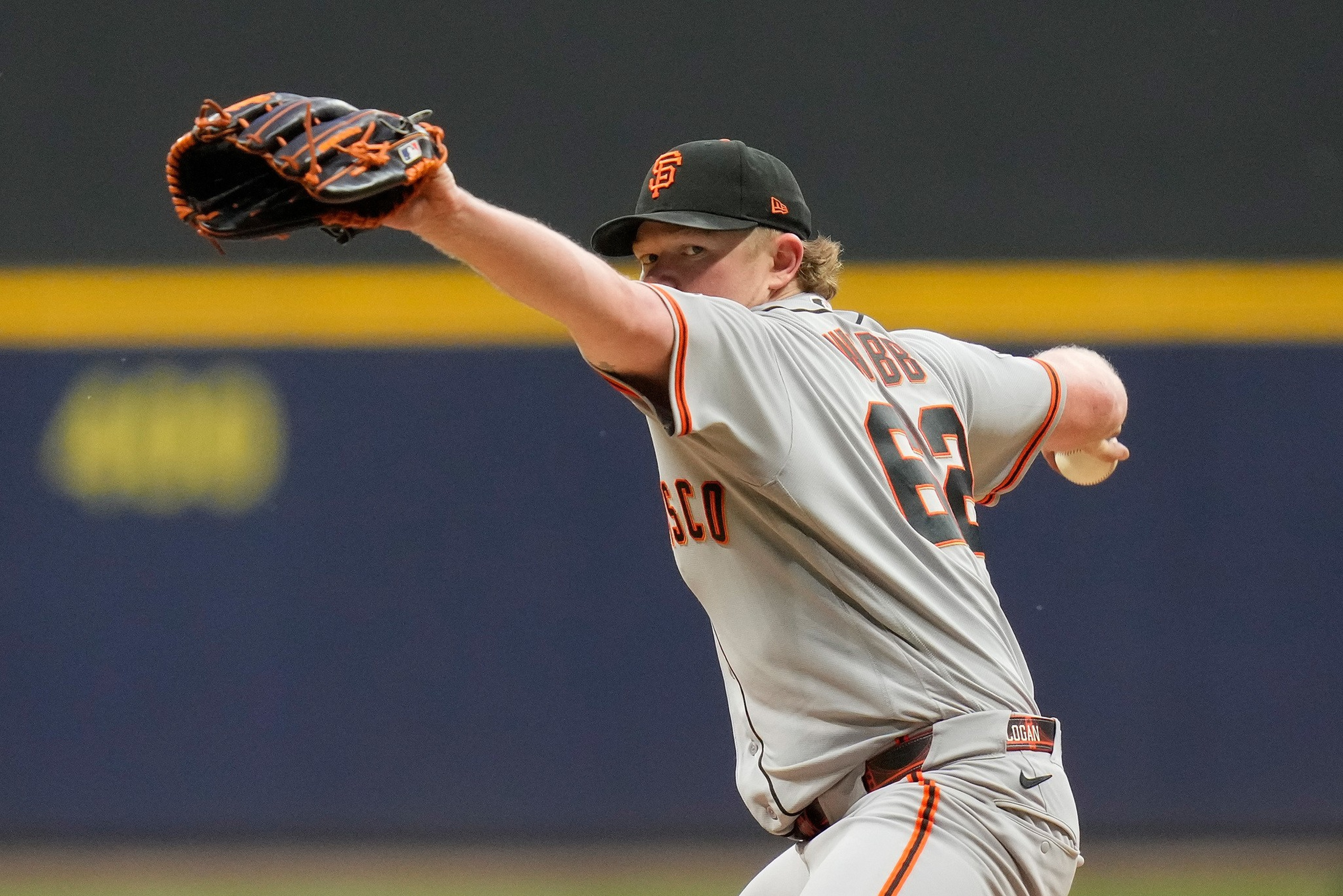 A baseball pitcher in a gray San Francisco Giants uniform is mid-throw, with his glove extended forward and the ball in his other hand.