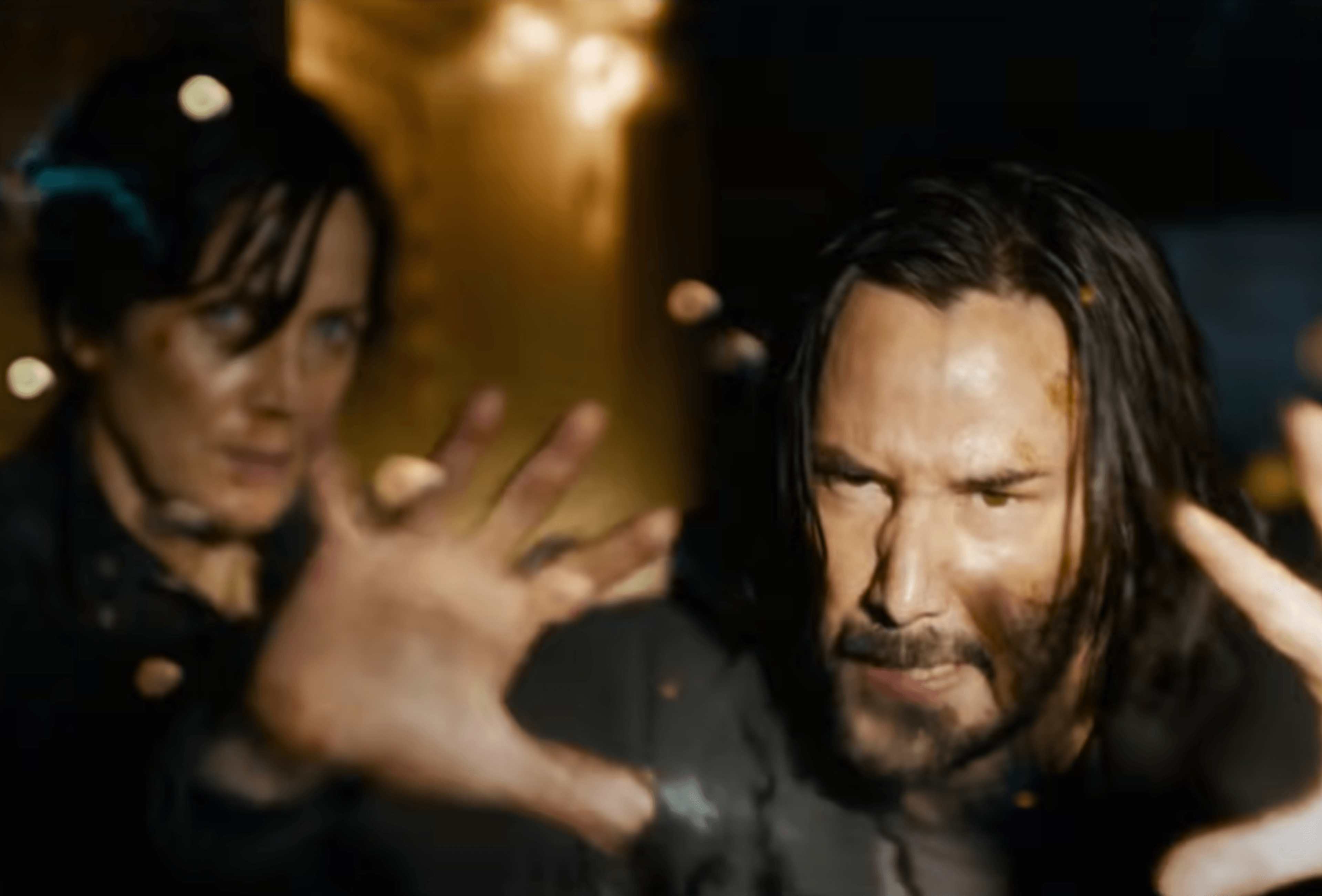 A man with long hair and a beard has his hands raised, looking intense, while a woman with short dark hair stands behind him in a dim setting.