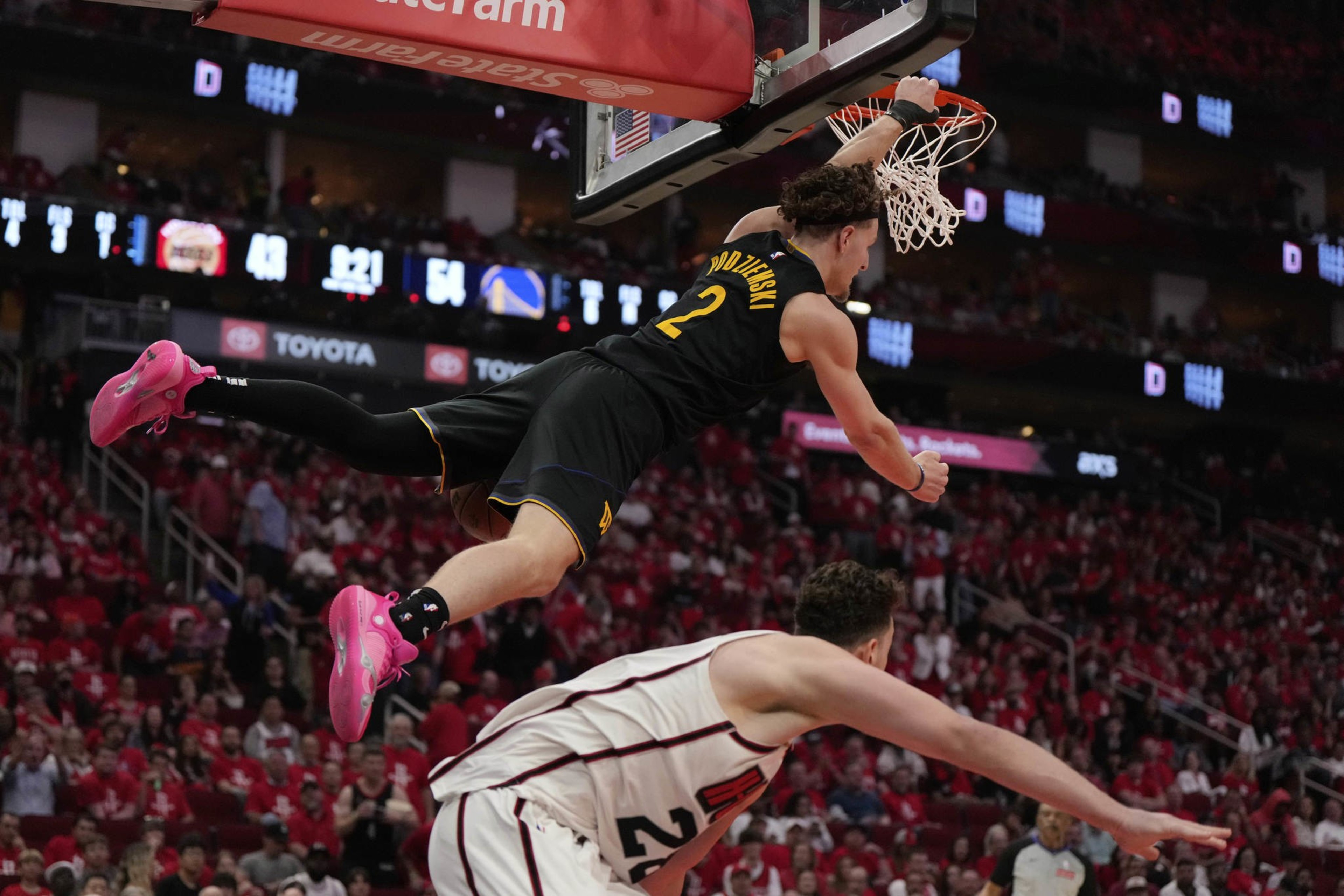 A basketball player in a black jersey, wearing bright pink shoes, is hanging on the rim after a dunk. A player in a white jersey stands below him.