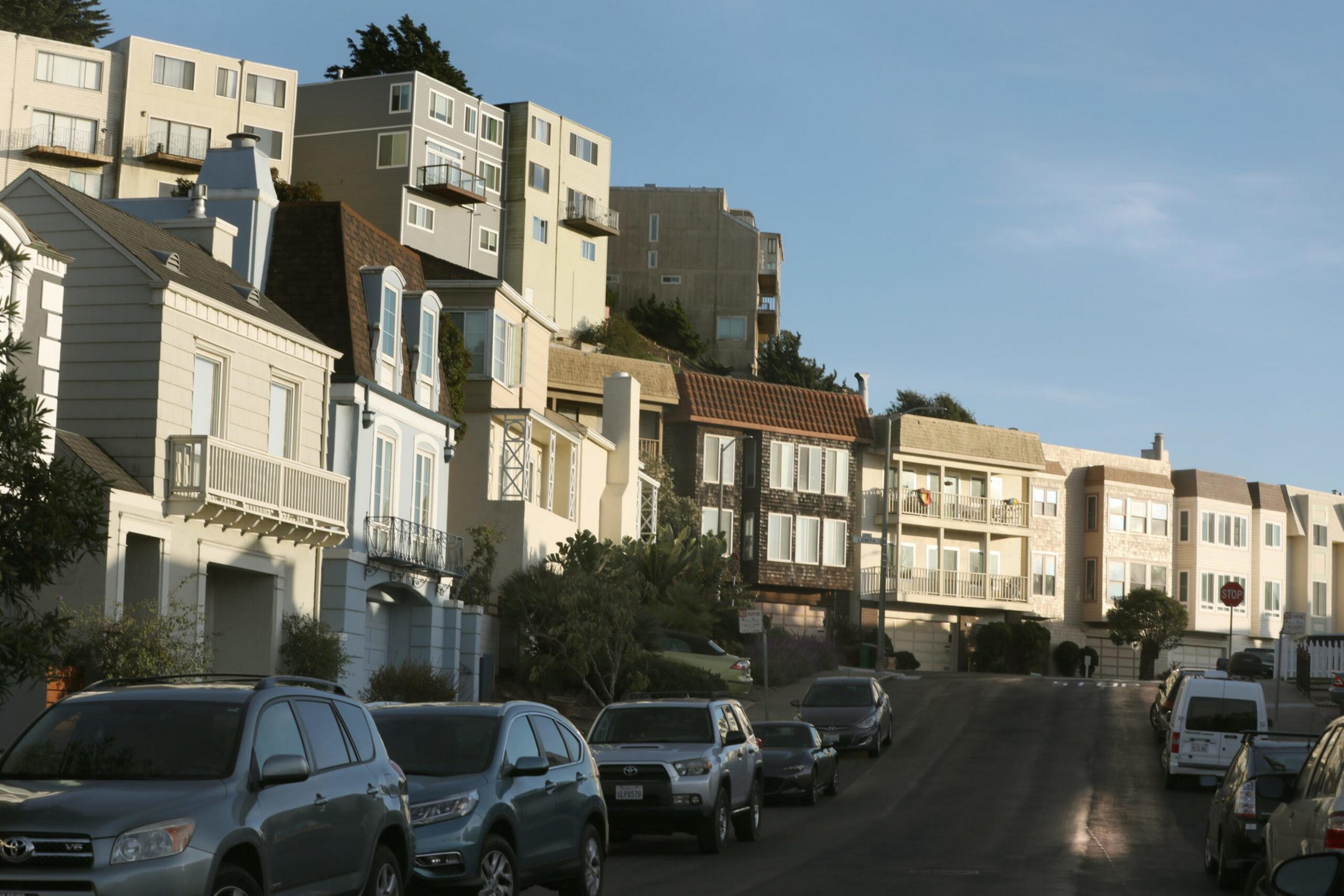 A row of multi-story houses with balconies lines a street filled with parked cars under a clear sky in a residential neighborhood.