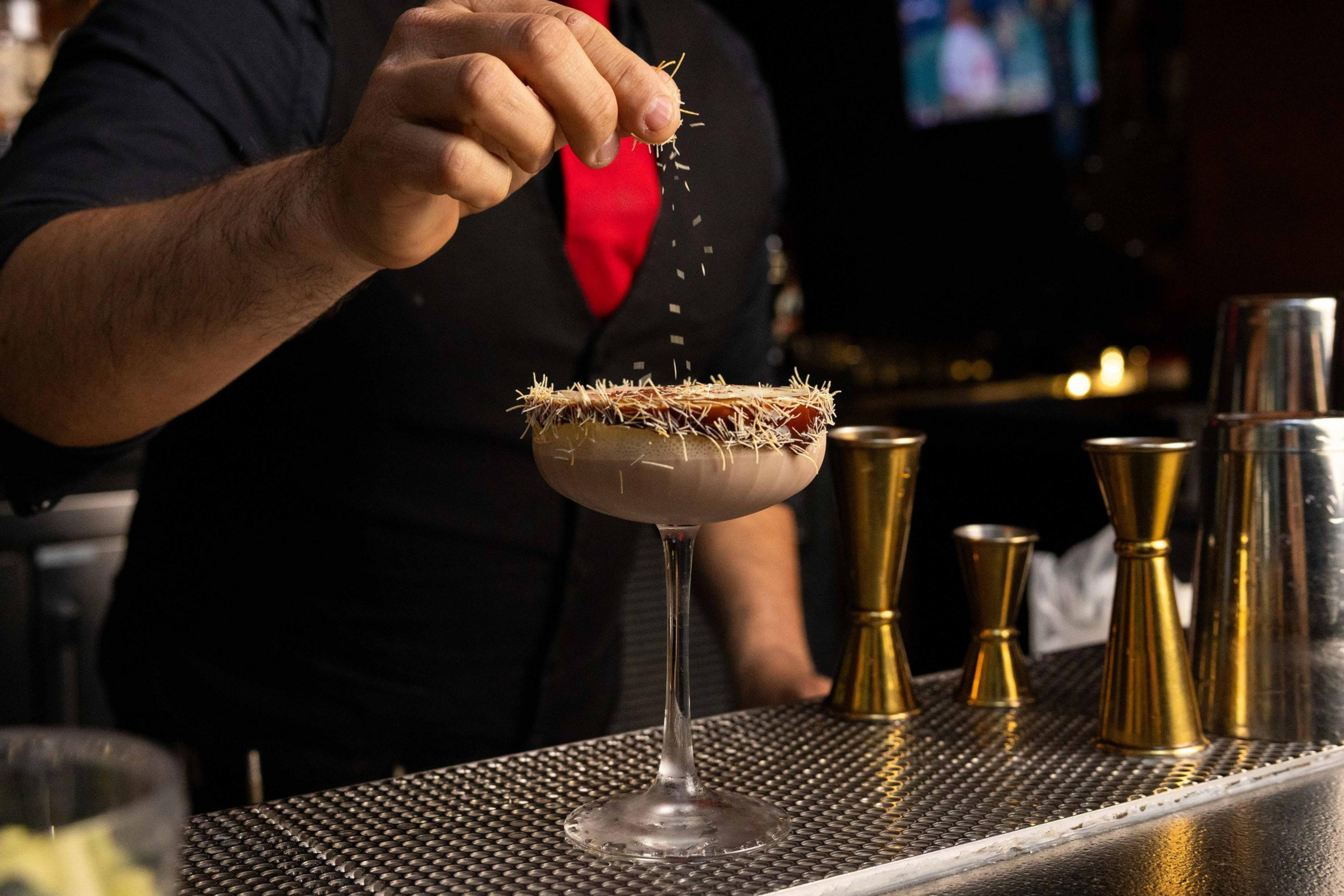A bartender sprinkles garnish onto a cocktail in a frosted glass with a decorated rim. The bar features golden measuring tools and soft ambient lighting.