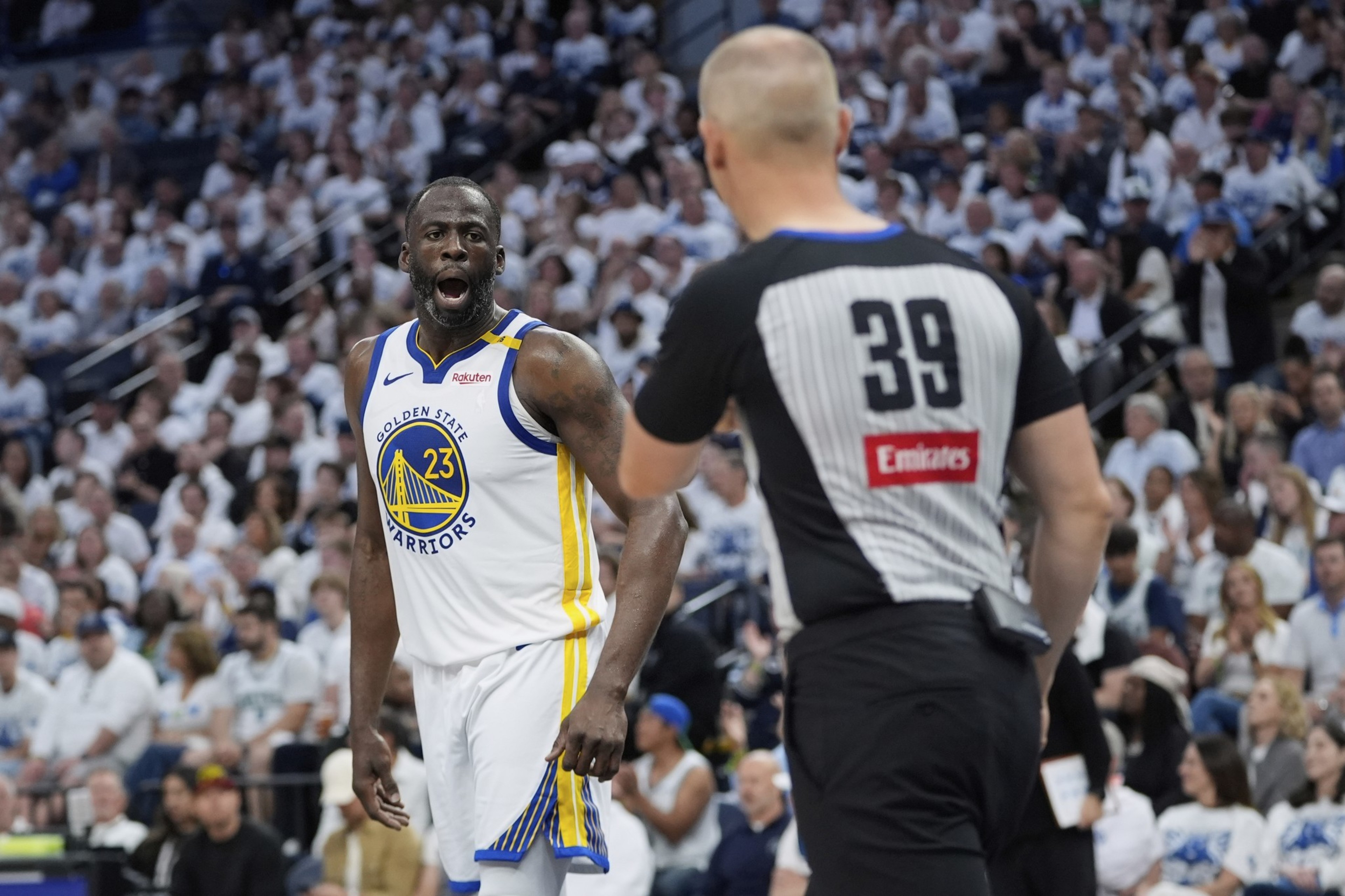 A basketball player in a white Golden State Warriors jersey is animatedly talking to a referee in a black-and-white striped shirt with "39" on the back.
