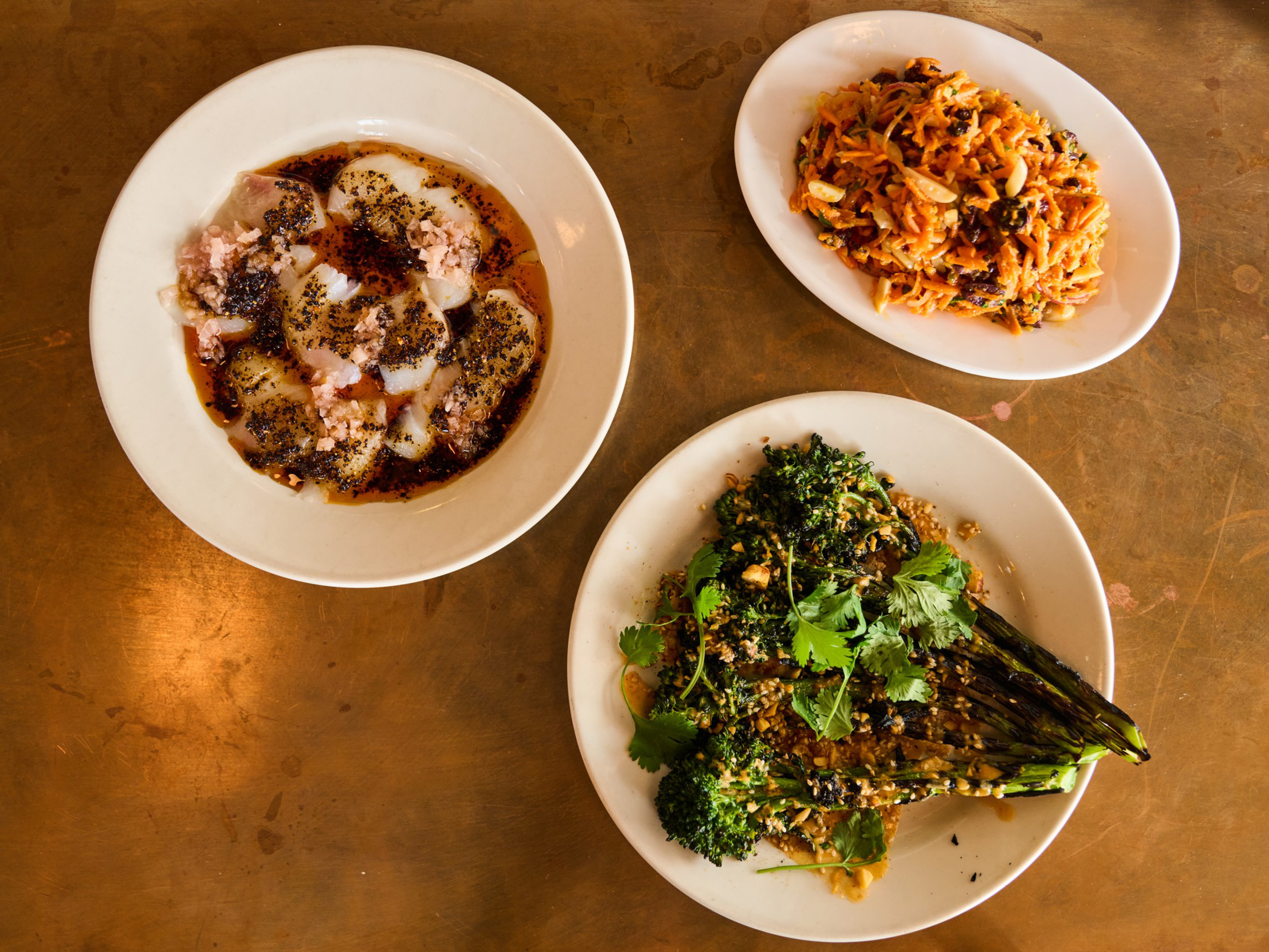Three dishes on a table: a plate of ravioli with sauce and cheese, a bowl of shredded carrot salad with raisins, and grilled broccoli topped with herbs.