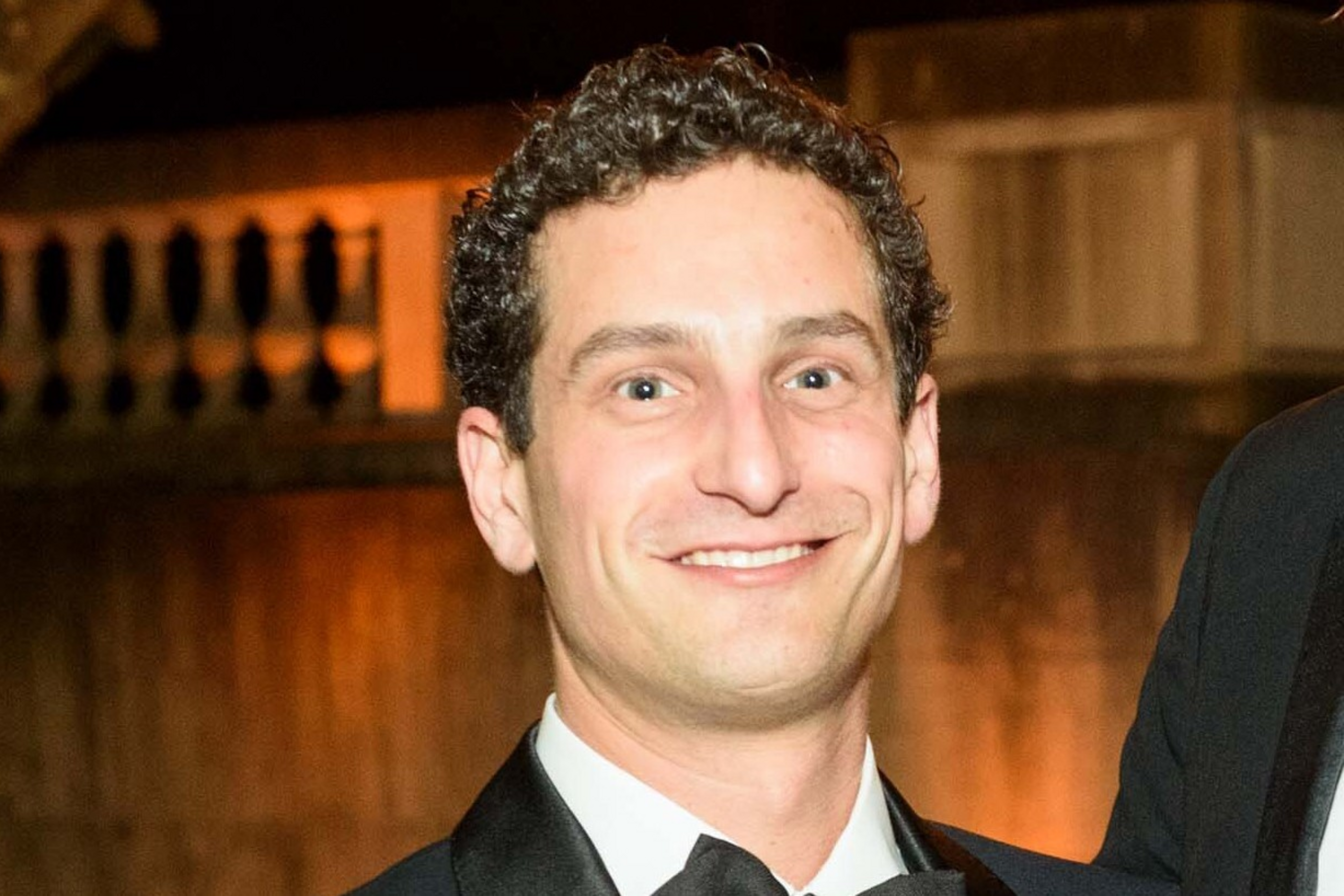 A man with curly dark hair is smiling, wearing a black tuxedo and bow tie against a warmly lit background.