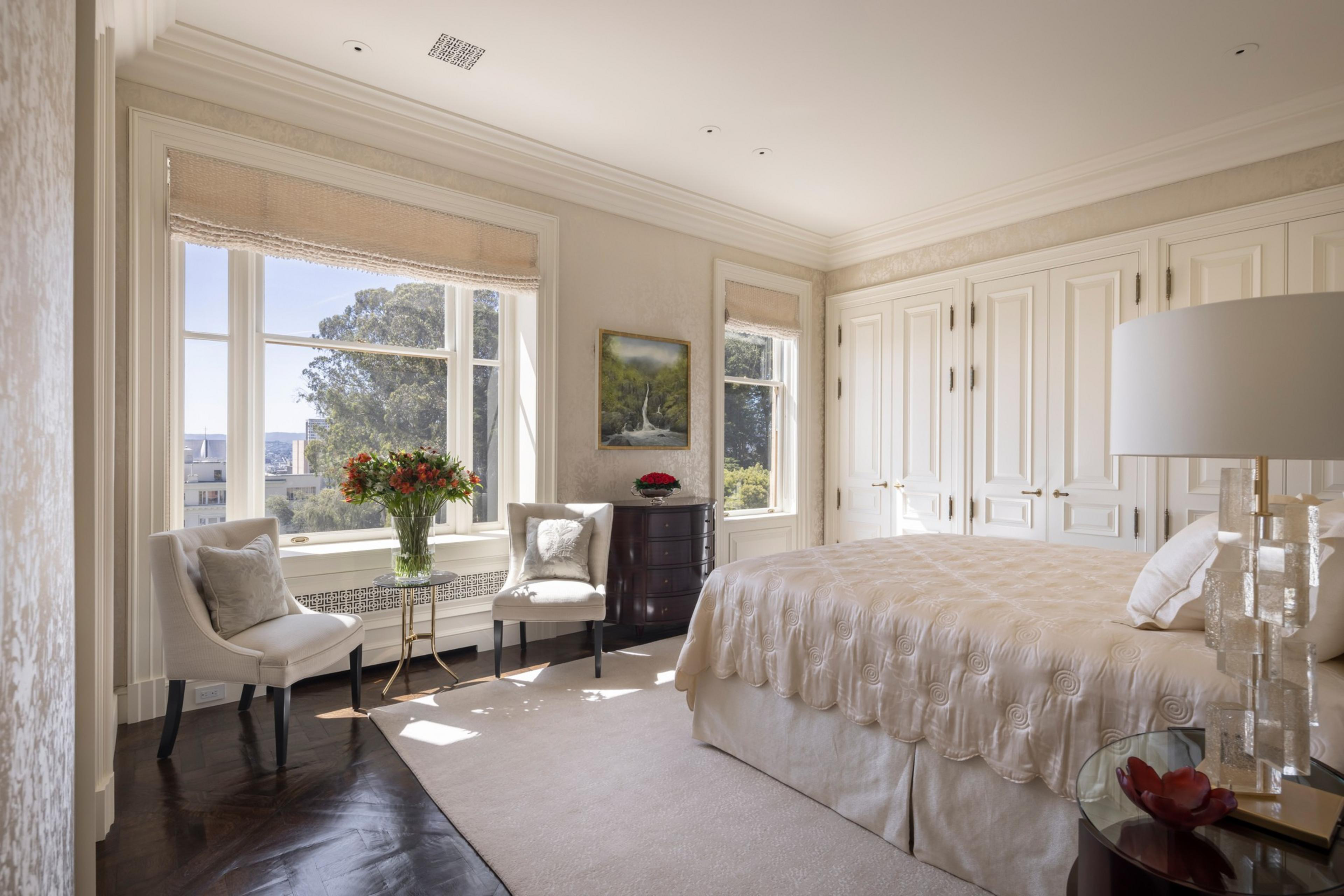 Two windows with chairs and a vase full of flowers nearby boast raised curtains allowing sunlight into a bedroom with a single large bed next to a small round end table. A bedroom boasts walk-in closets, sun-filled windows and dark wood floors at a San Francisco penthouse.