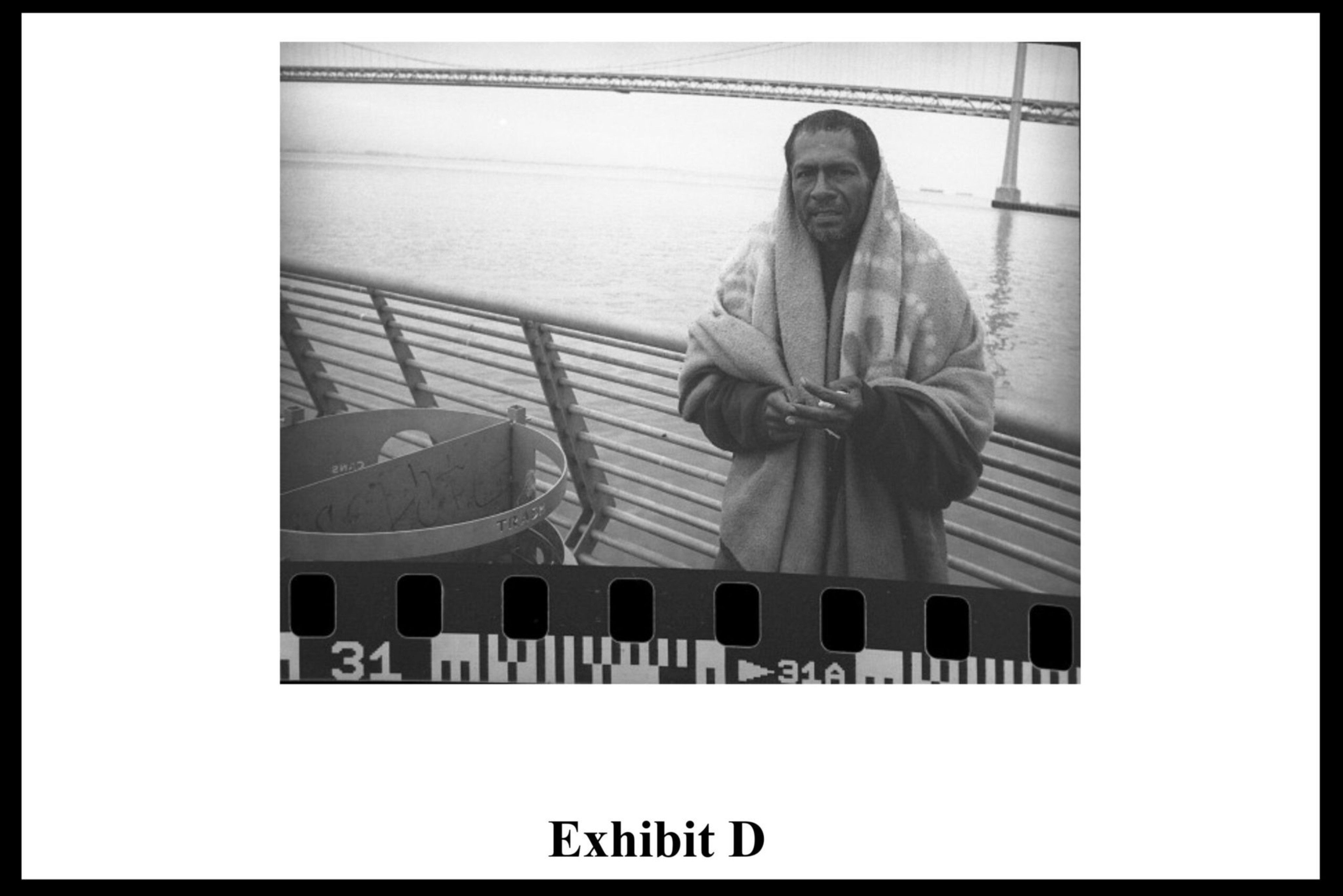 A man wrapped in a blanket stands near a metal railing by the water with a bridge in the background and a trash bin nearby.