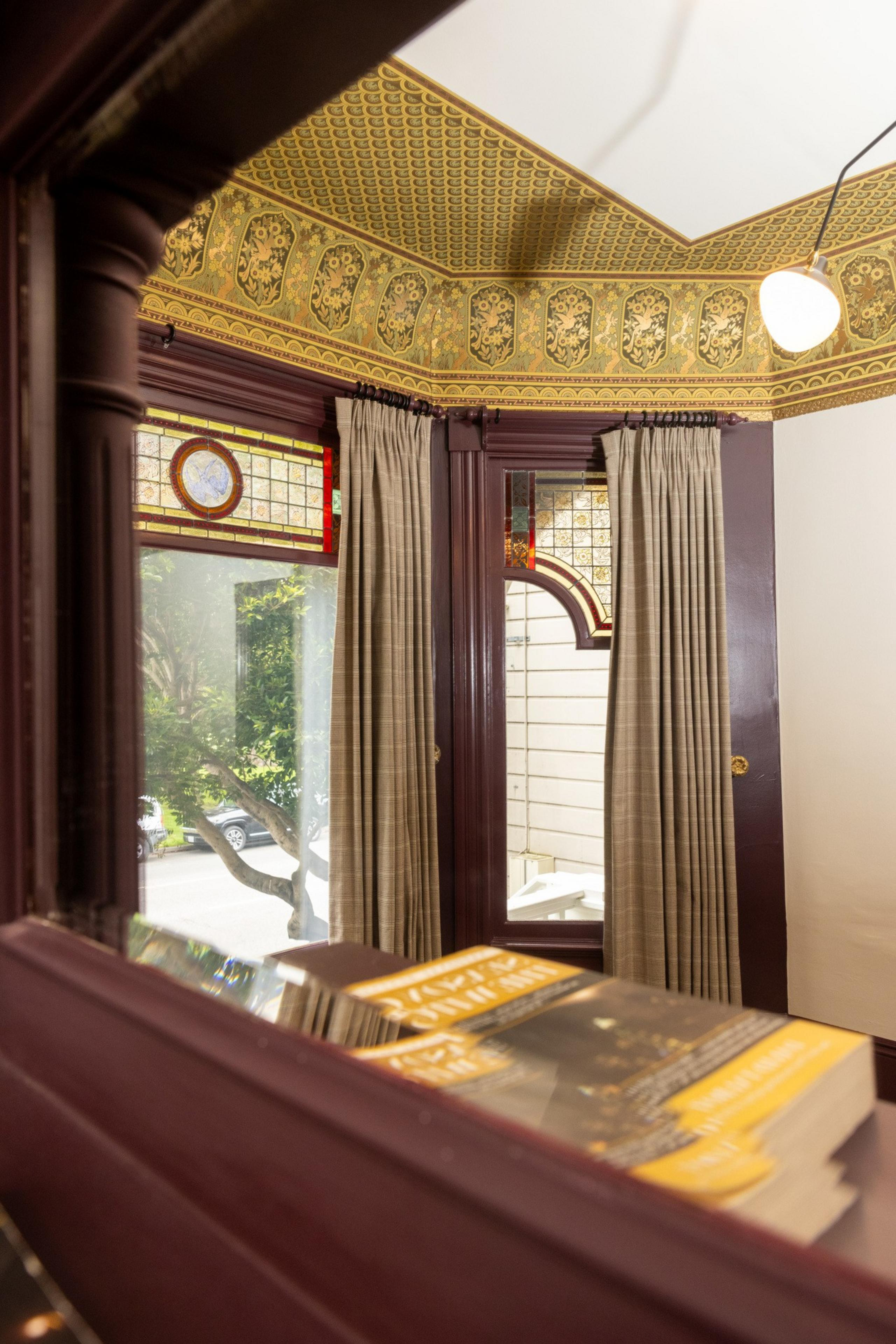 A room with ornate yellow ceiling moldings, large stained-glass windows, light curtains, and a reflective surface showing a stack of books in the foreground.