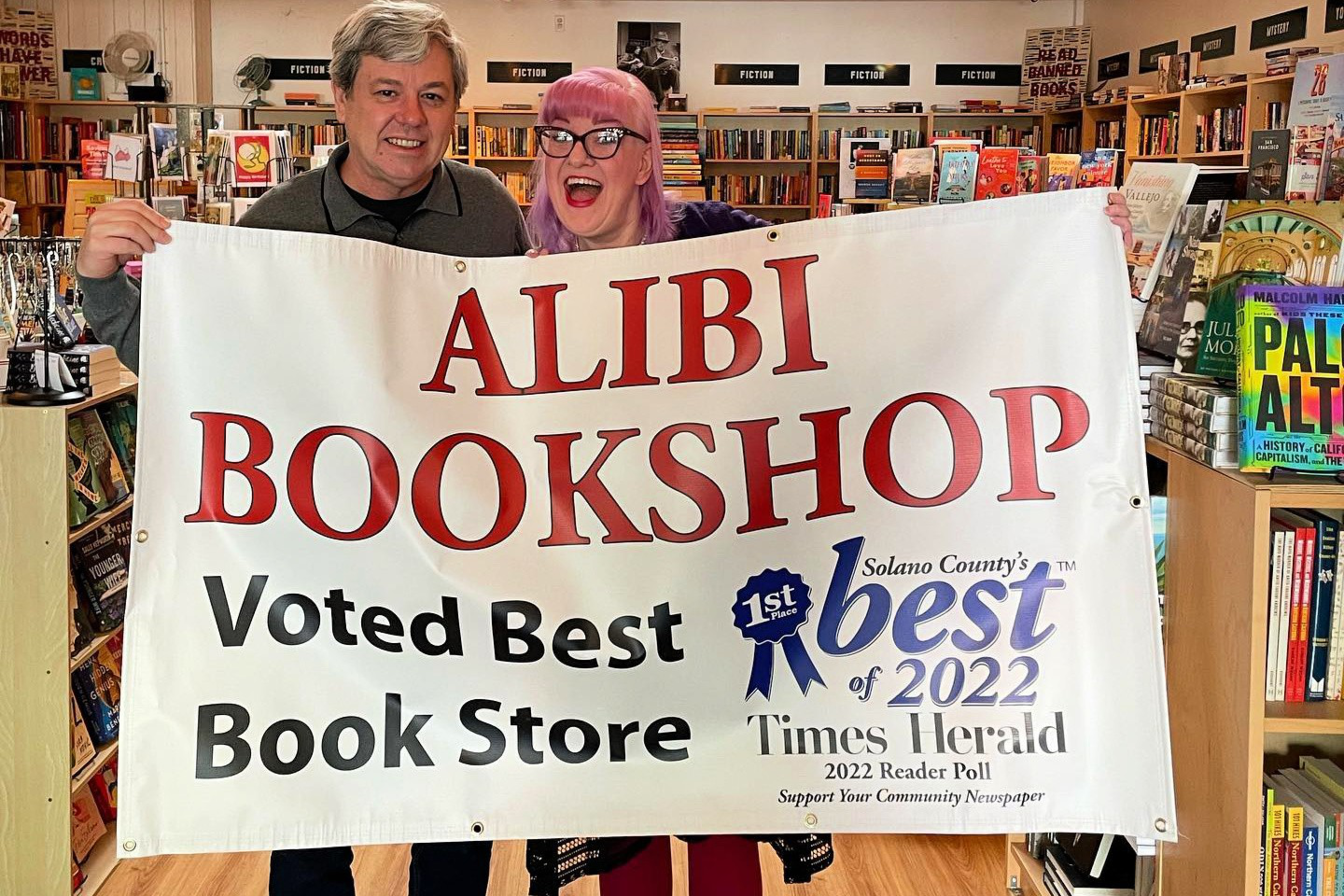Two people holding a sign saying Alibi Bookshop. 