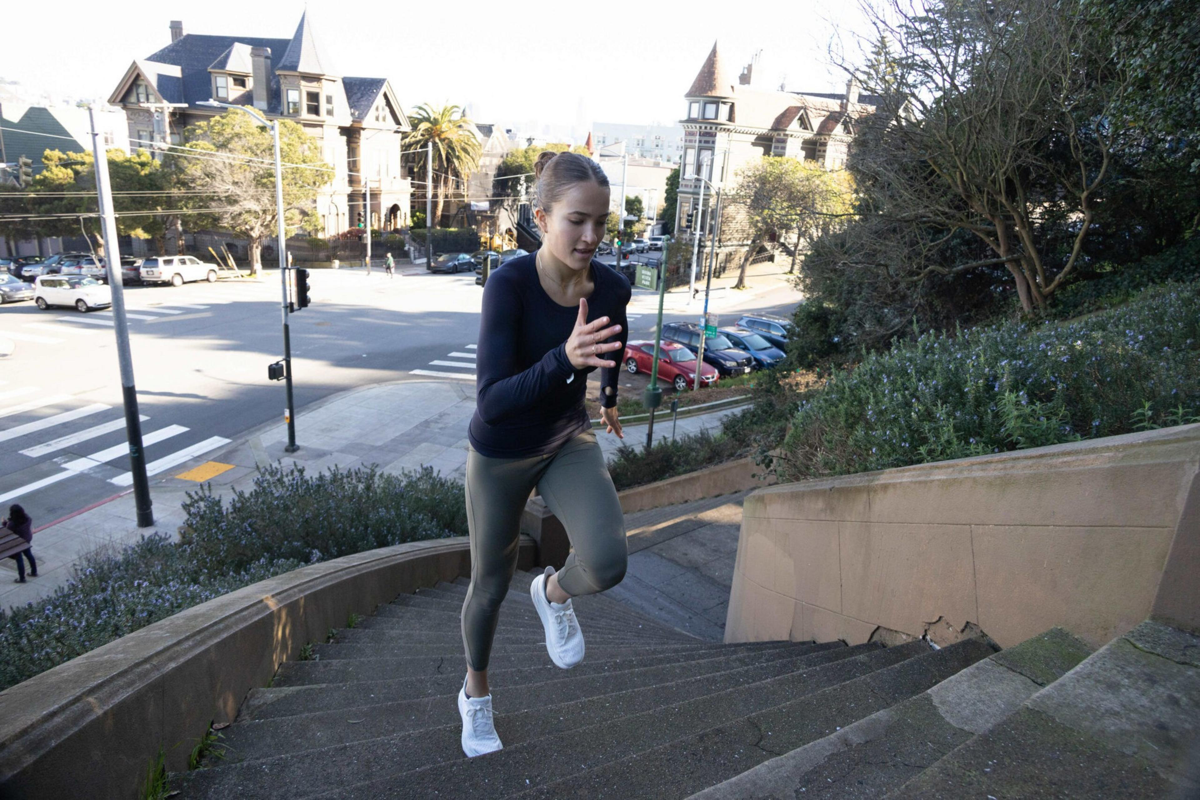 A woman in athletic wear runs up outdoor stone stairs with houses, cars, and trees visible in the background on a sunny day.