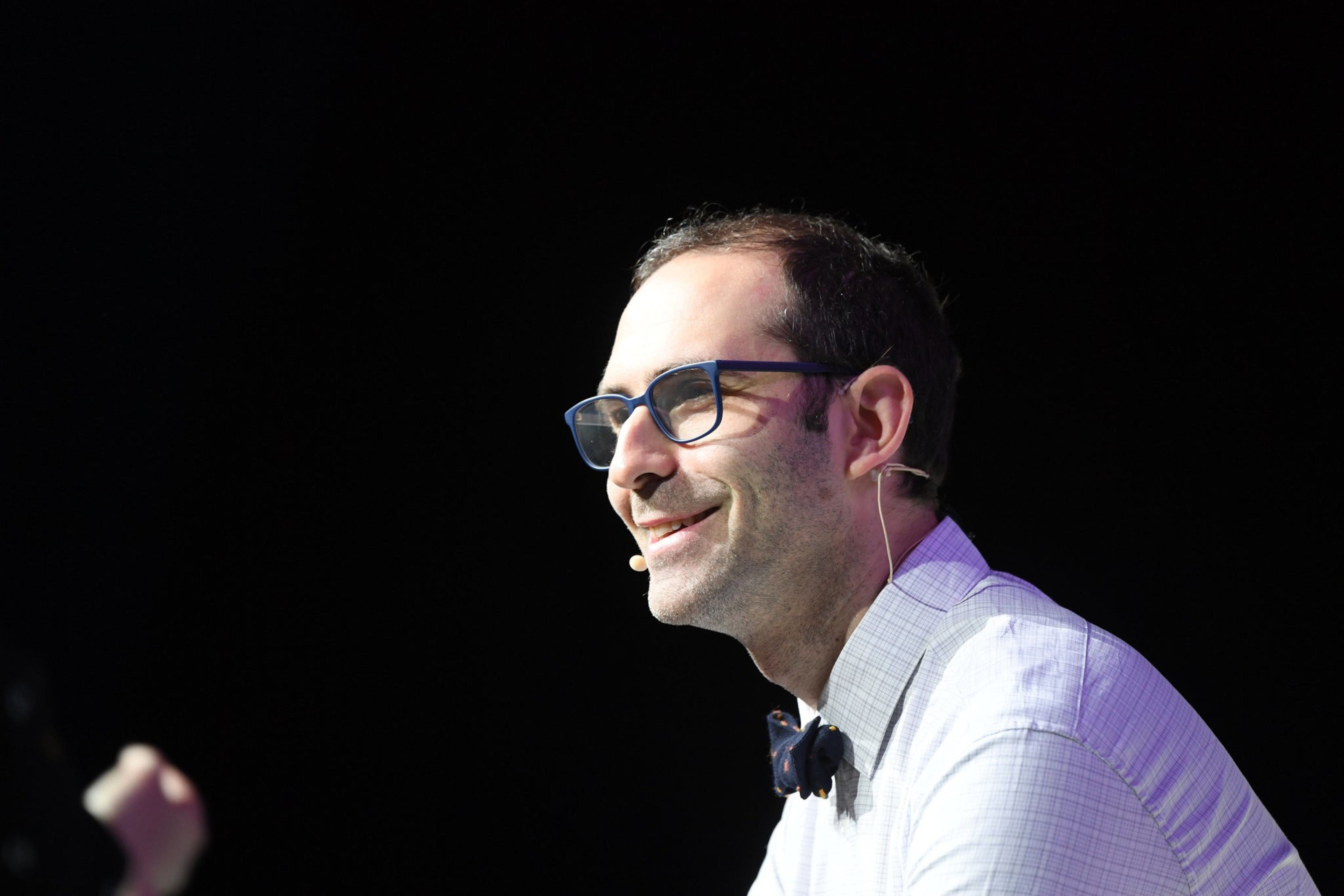 A man with blue-framed glasses smiles against a black background.