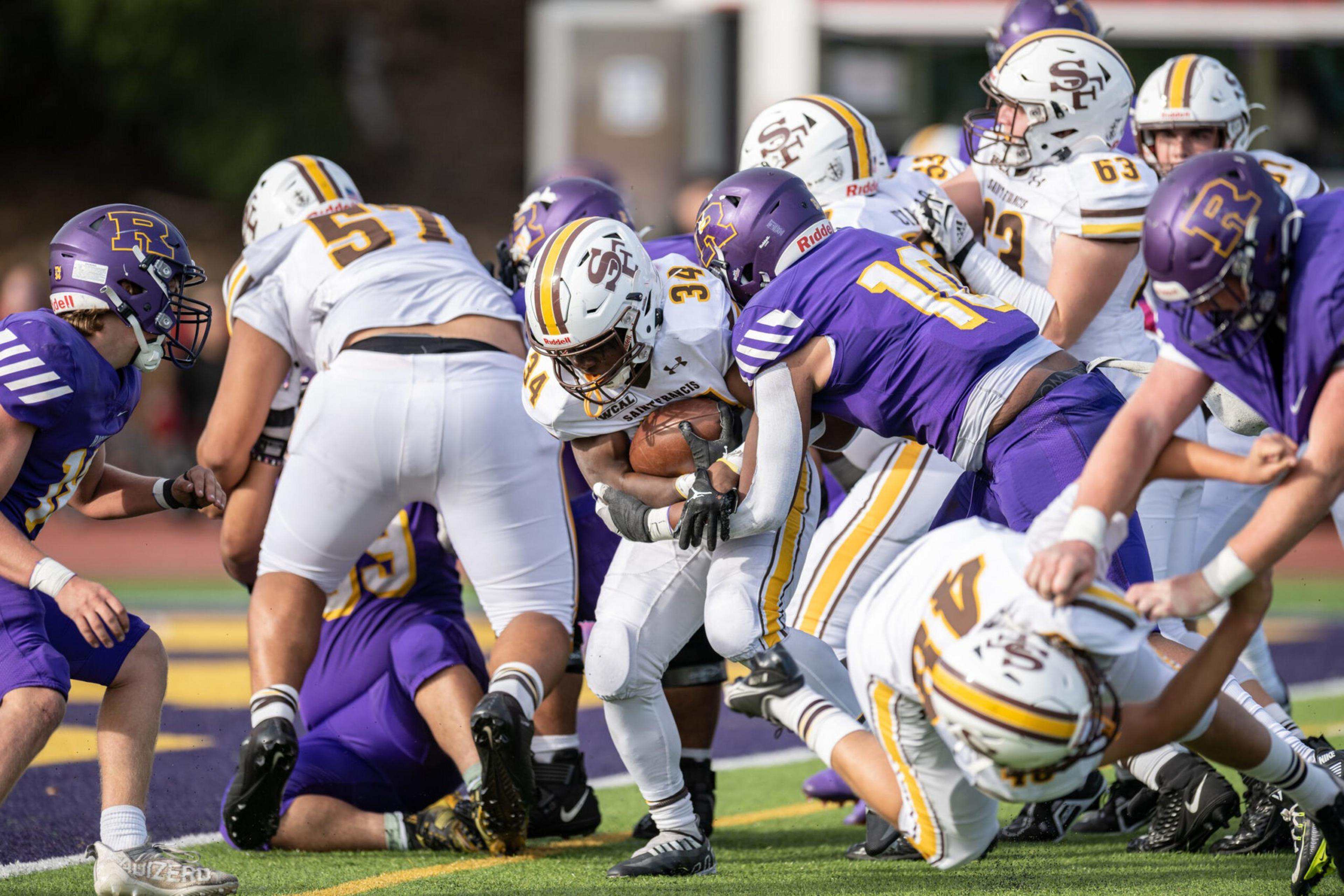 A football player in a white uniform is running with the ball while being tackled by two defenders in purple uniforms during a game.