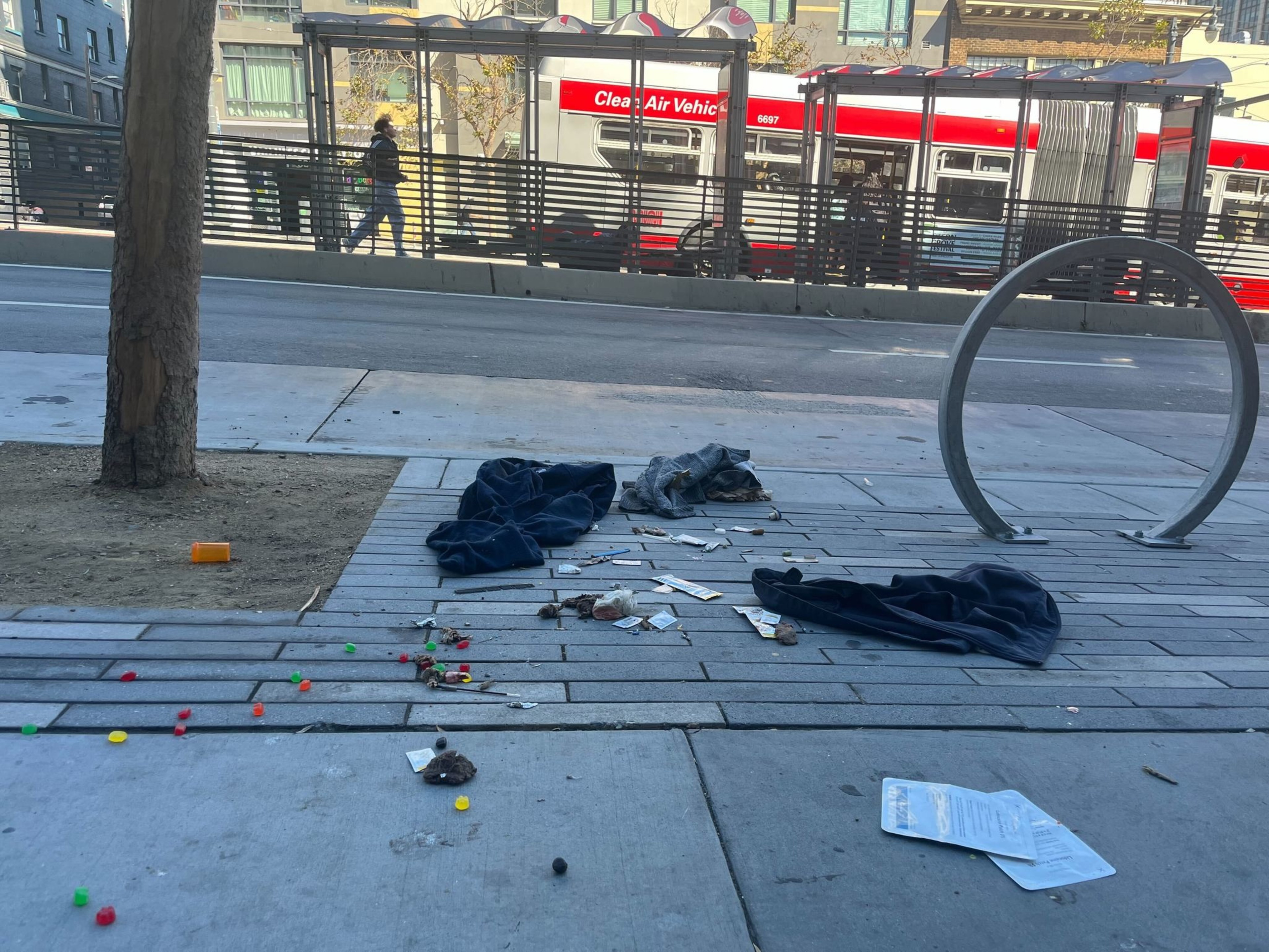 There are discarded clothes, candy, and litter on a sidewalk near a tree and a bike rack. Across the street, pedestrians walk by and a red bus is seen.