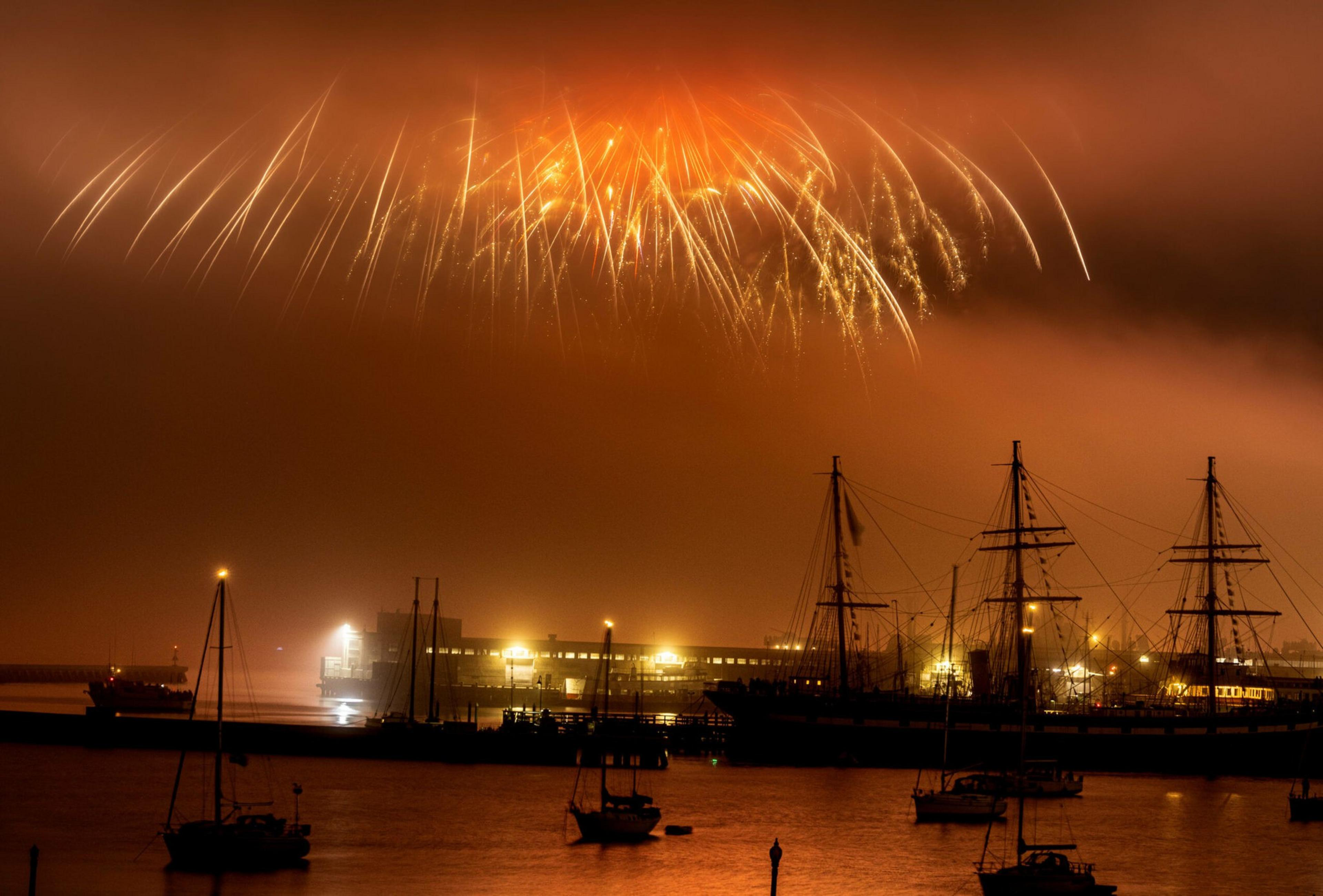 Golden fireworks burst low and wide over a harbor filled with silhouetted sailboats and a brightly lit dock at night.