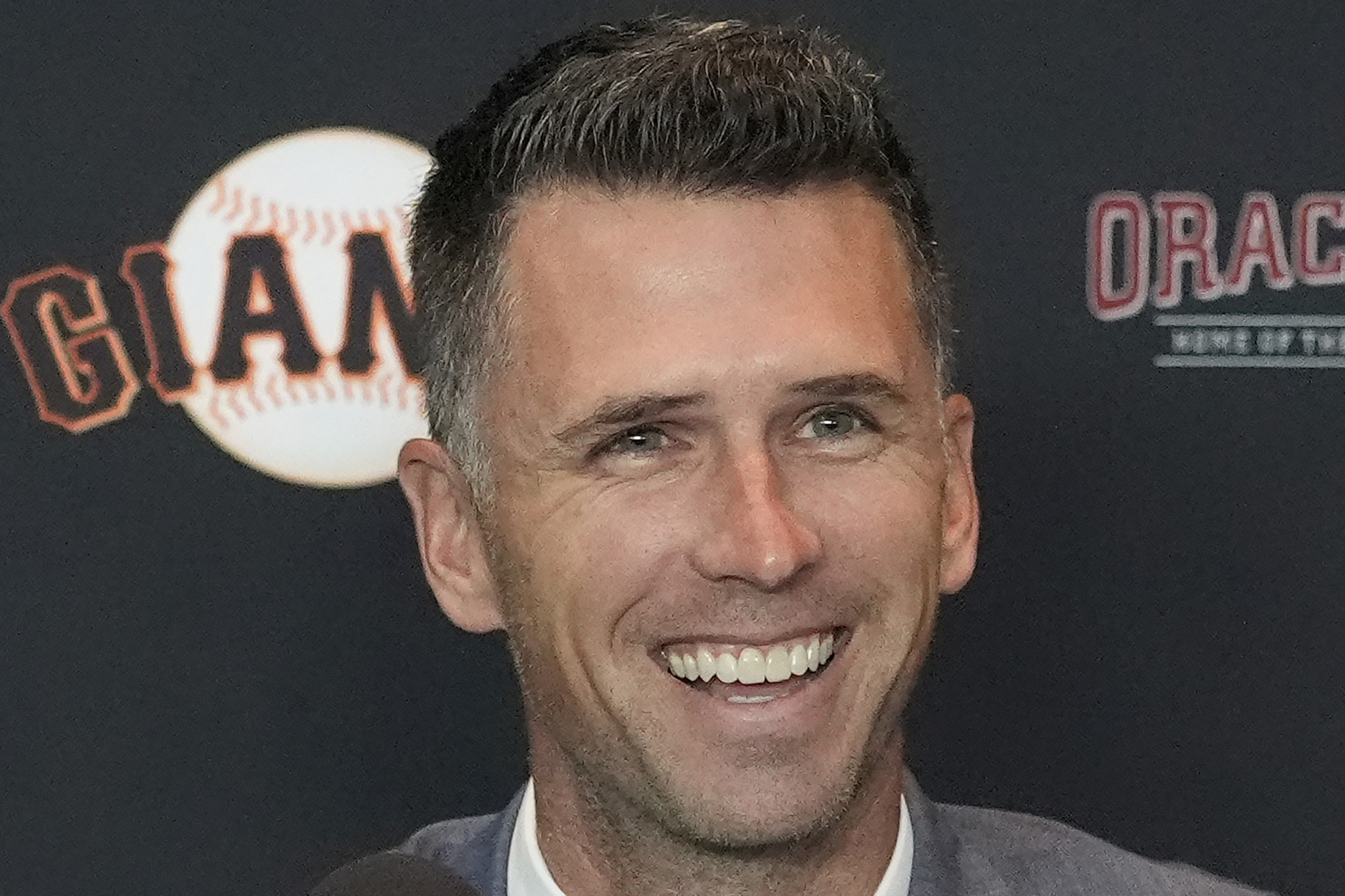A smiling man in a suit is speaking at a press event with "Giants" and "Oracle" logos in the background. A microphone with the team logo is in front of him.
