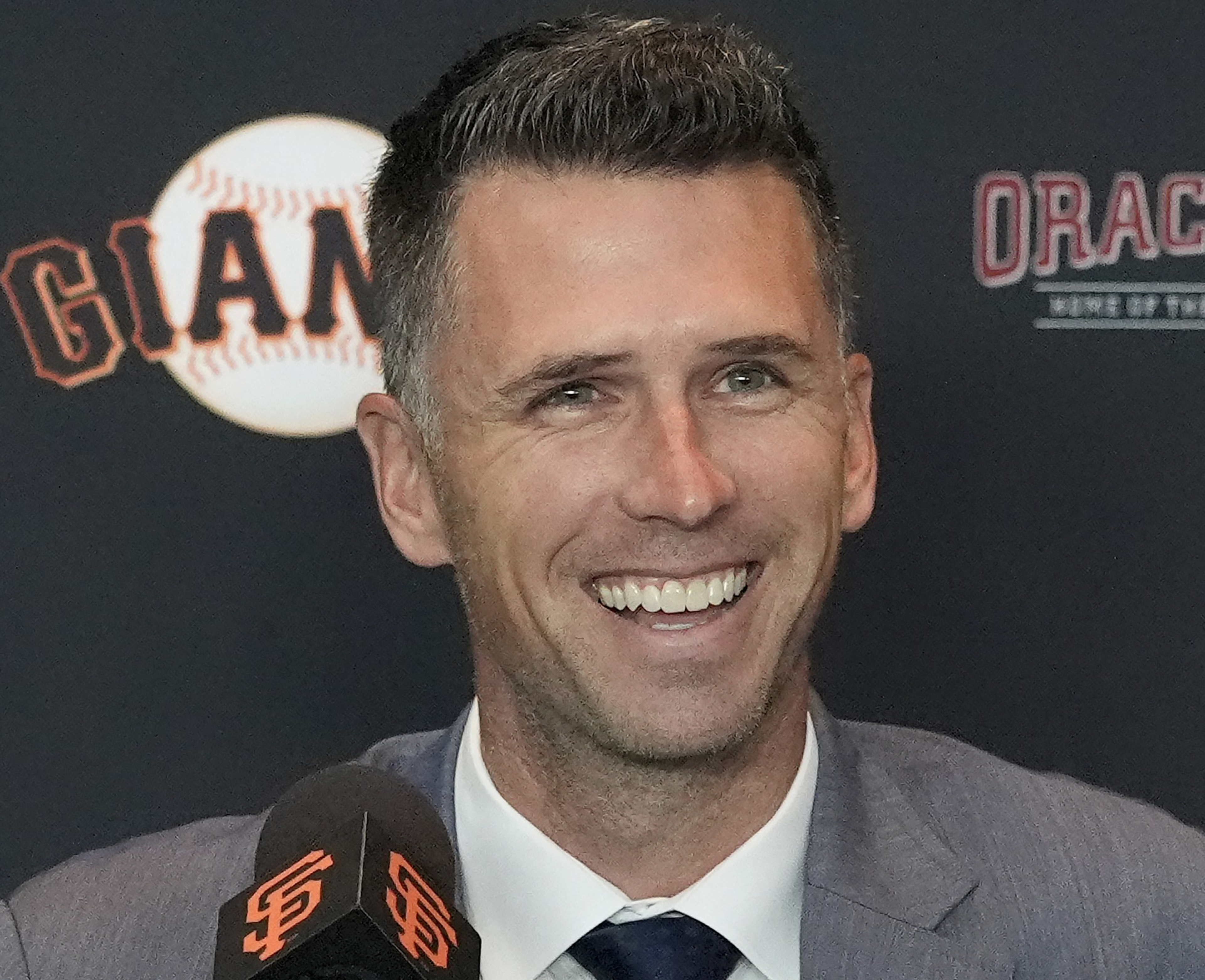 A smiling man in a suit is speaking at a press event with "Giants" and "Oracle" logos in the background. A microphone with the team logo is in front of him.