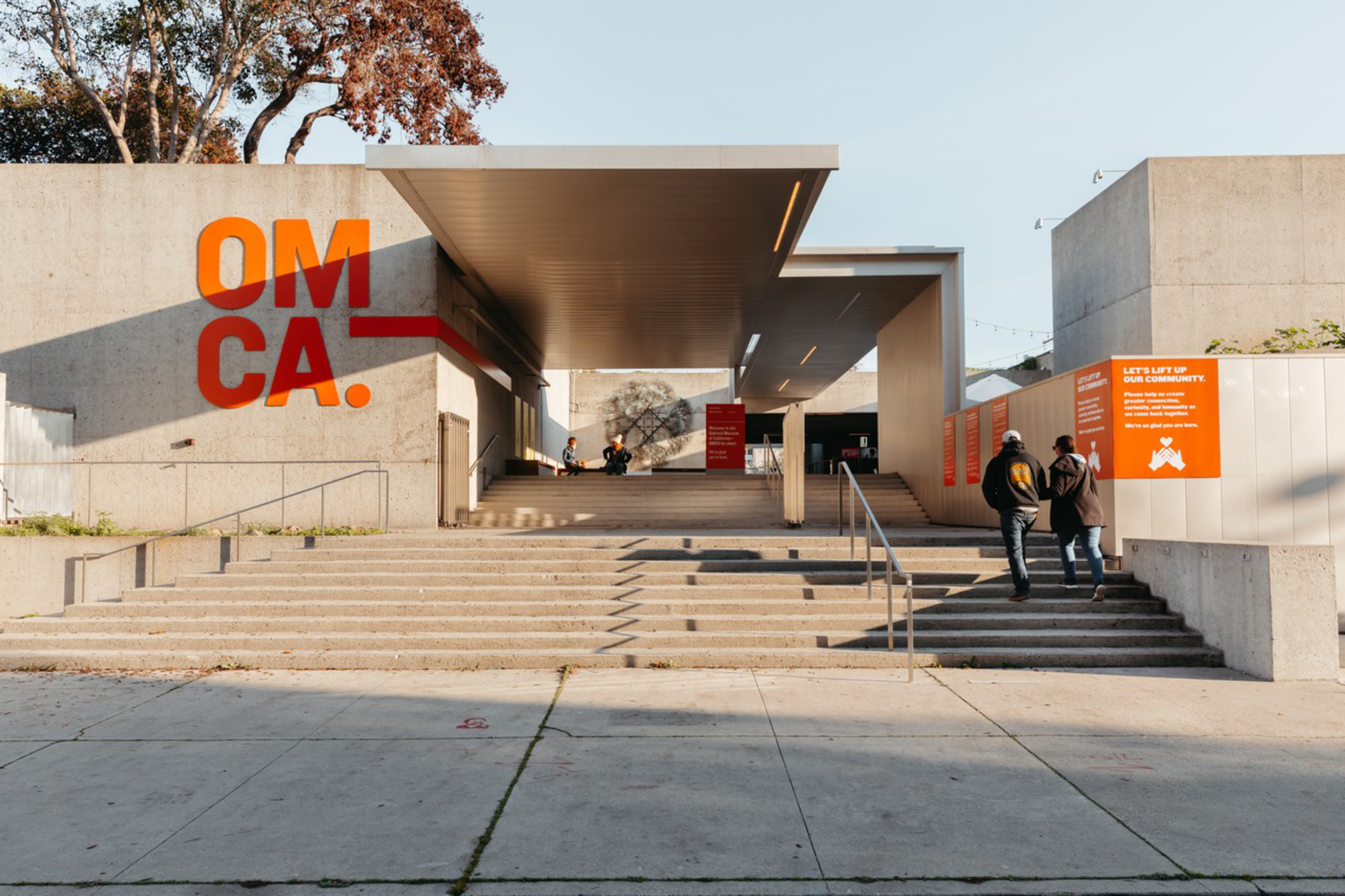 A modern museum entrance with wide stairs, bold orange “OMCA.” letters on the wall, two people walking up, and others sitting inside.