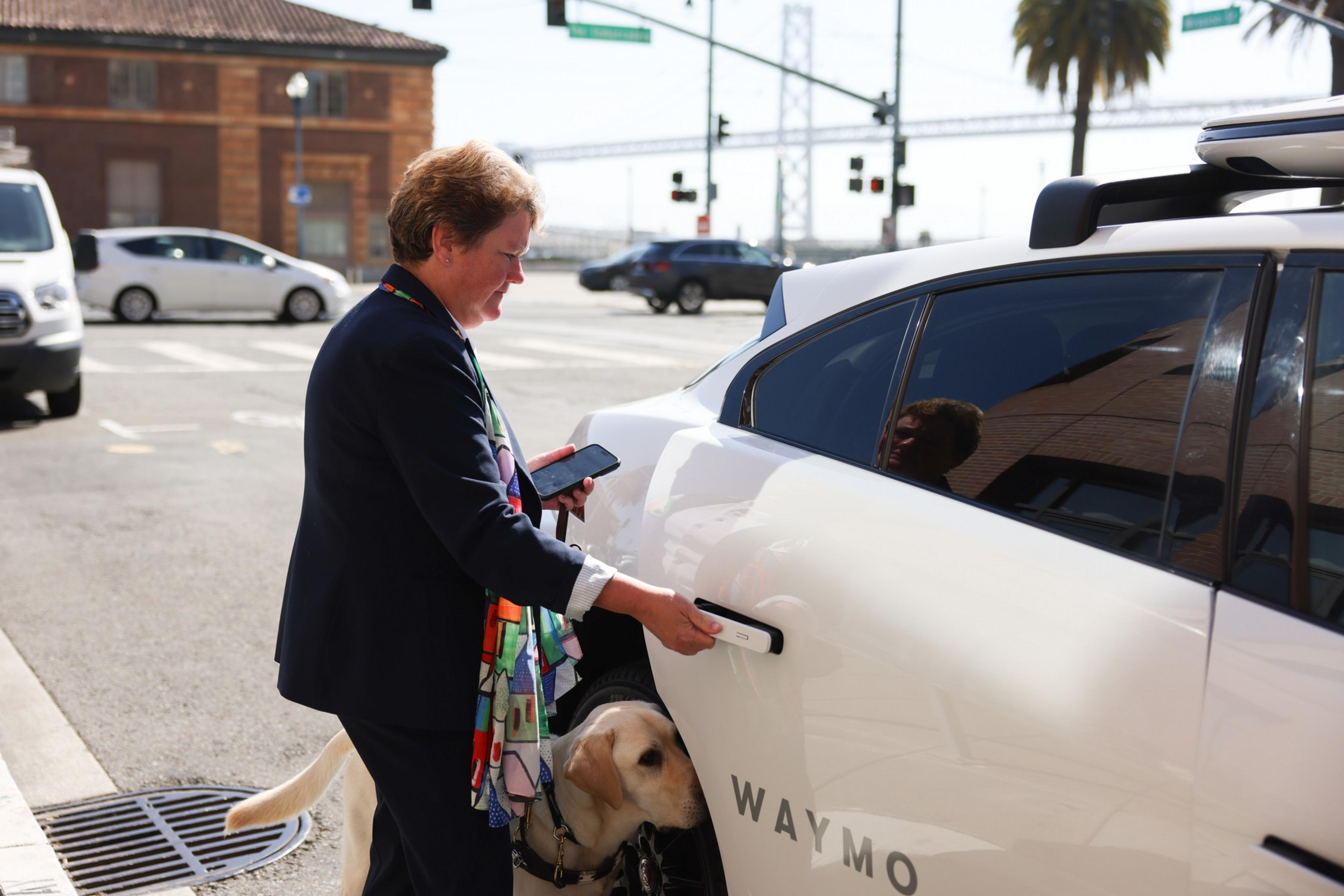 A person opens a white Waymo car door while holding a phone, accompanied by a guide dog near an urban street intersection.