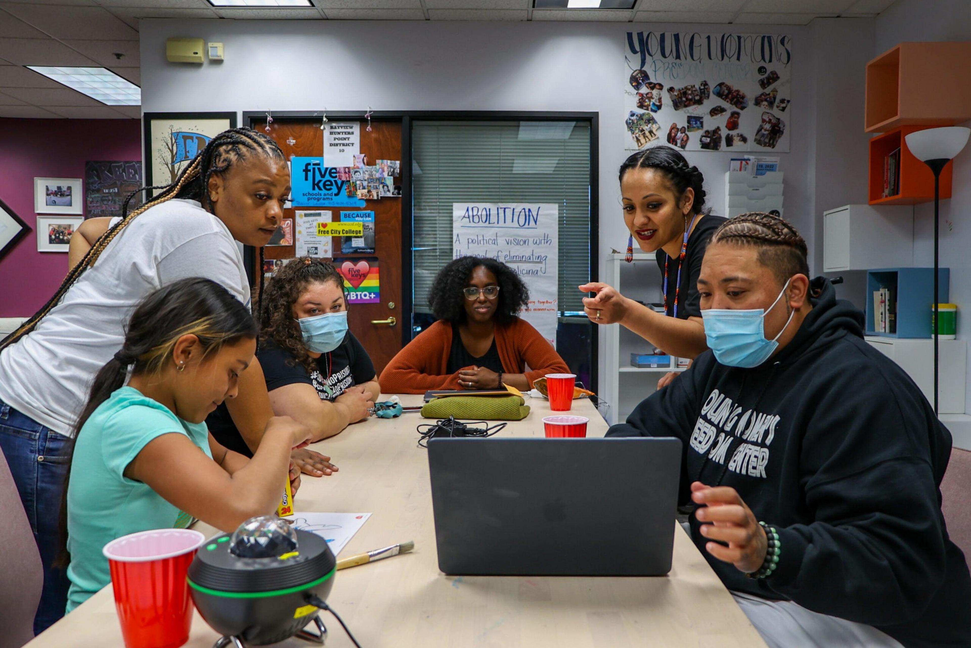 Six people engage around a laptop at a table in a room with colorful posters and red cups, two of them wearing face masks.