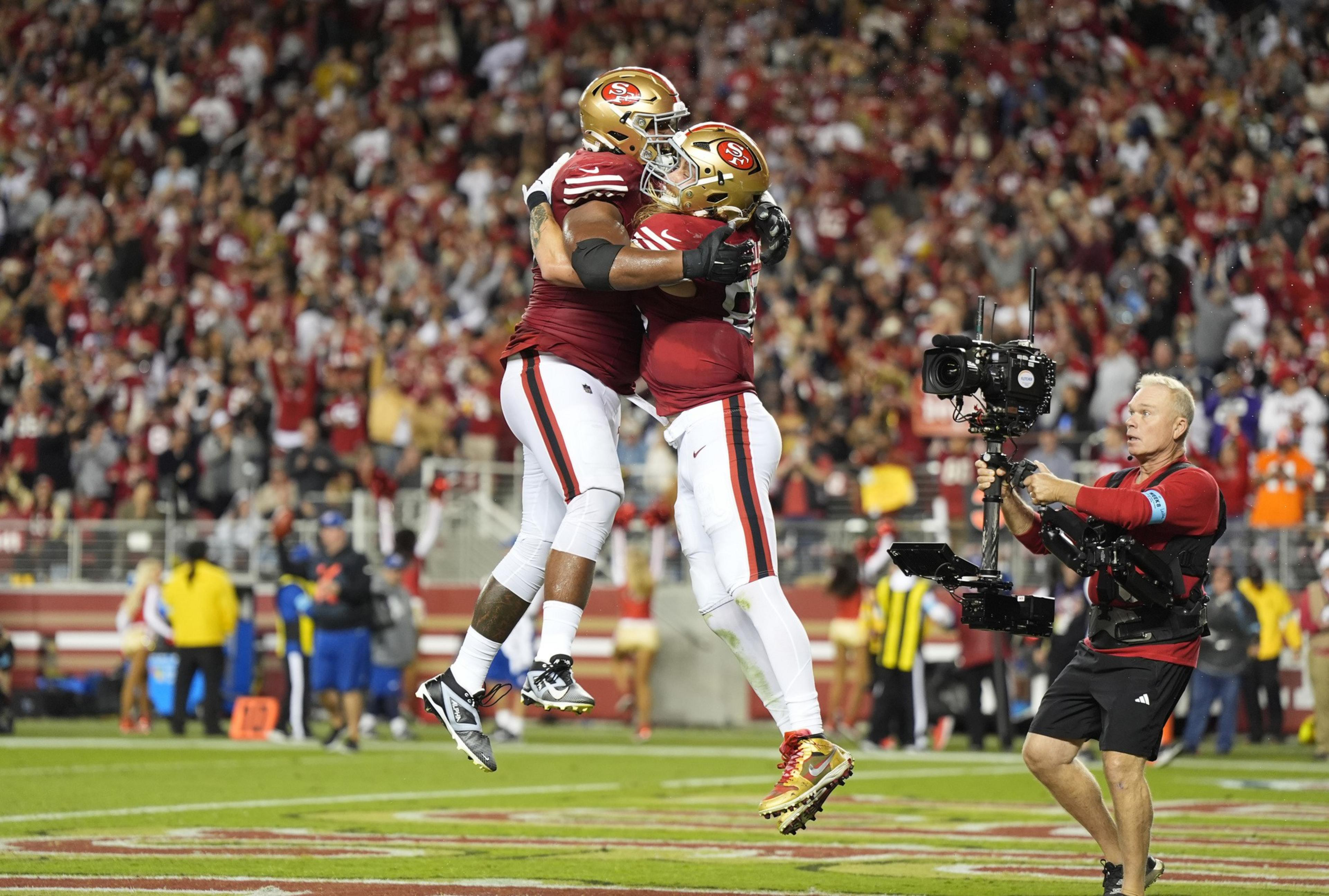 Two football players in red uniforms joyfully jump and embrace on the field, with a cameraman capturing the moment and a cheering crowd in the background.