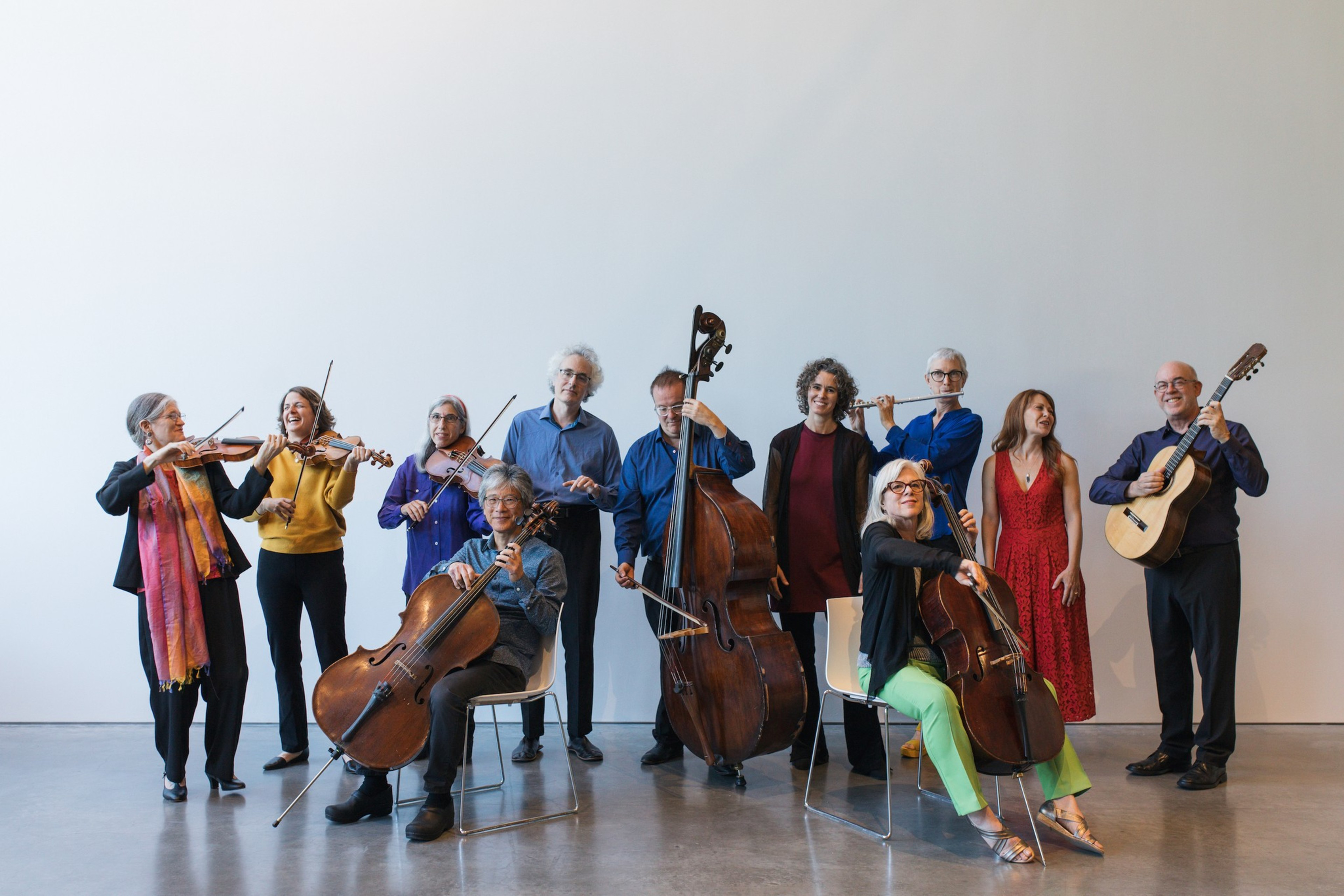 A diverse group of musicians plays various instruments, including violins, cellos, a double bass, a flute, and a guitar, against a plain white wall.