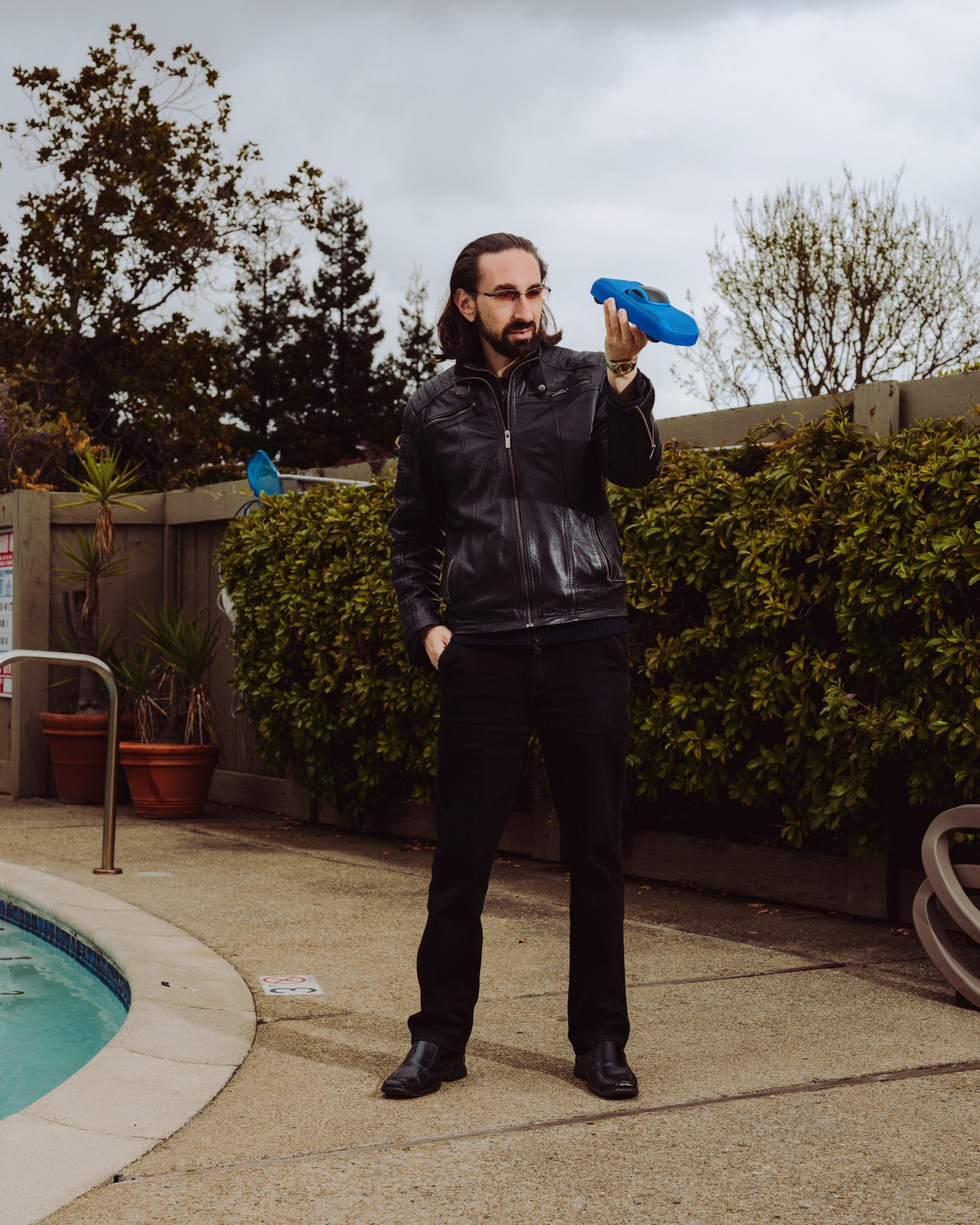 A man in a black leather jacket stands by a pool, holding a blue object. The background features trees, cloudy skies, and a hedge.
