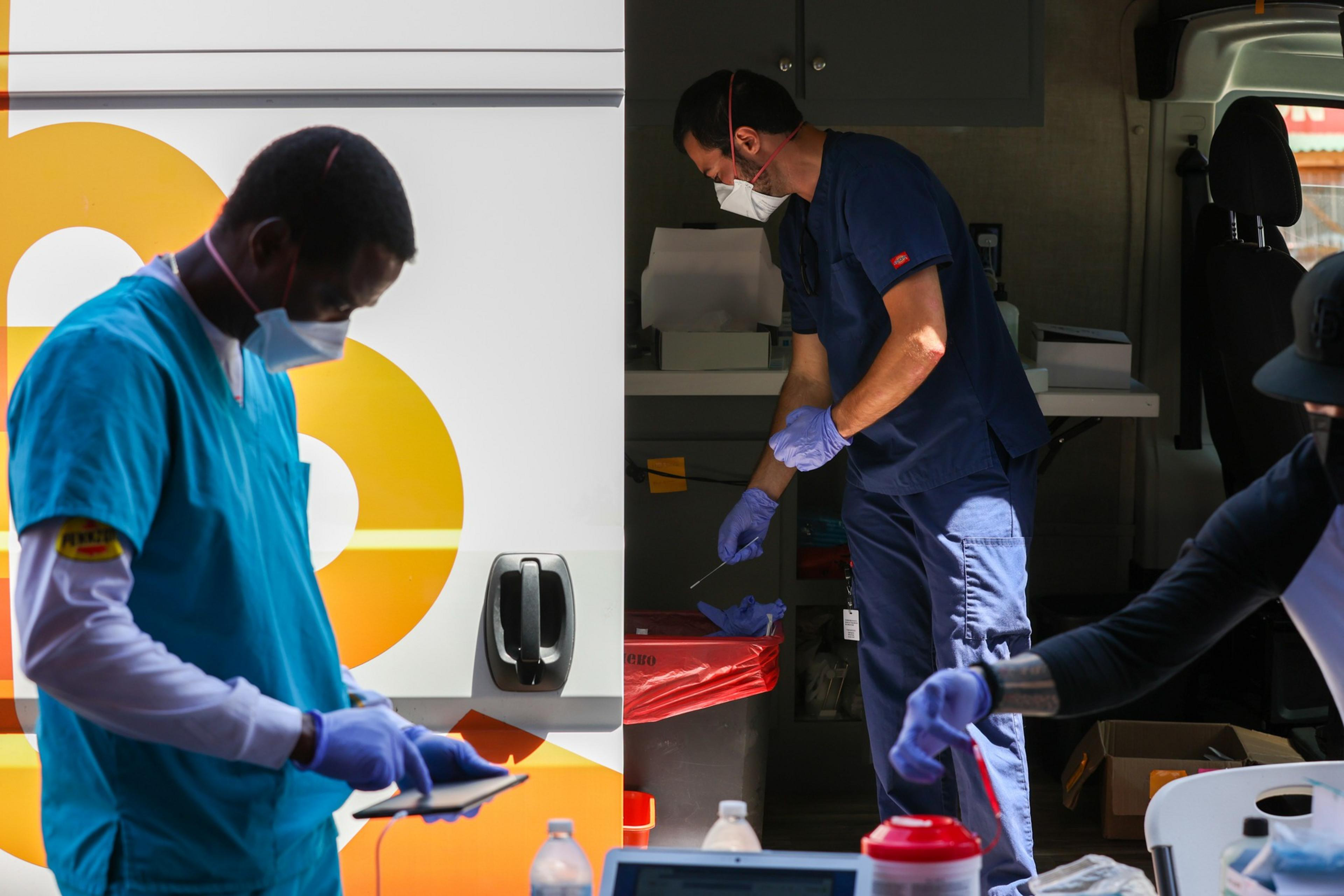 Two healthcare workers in masks and gloves are engaged in a mobile medical testing or vaccination setup near a vehicle with equipment.