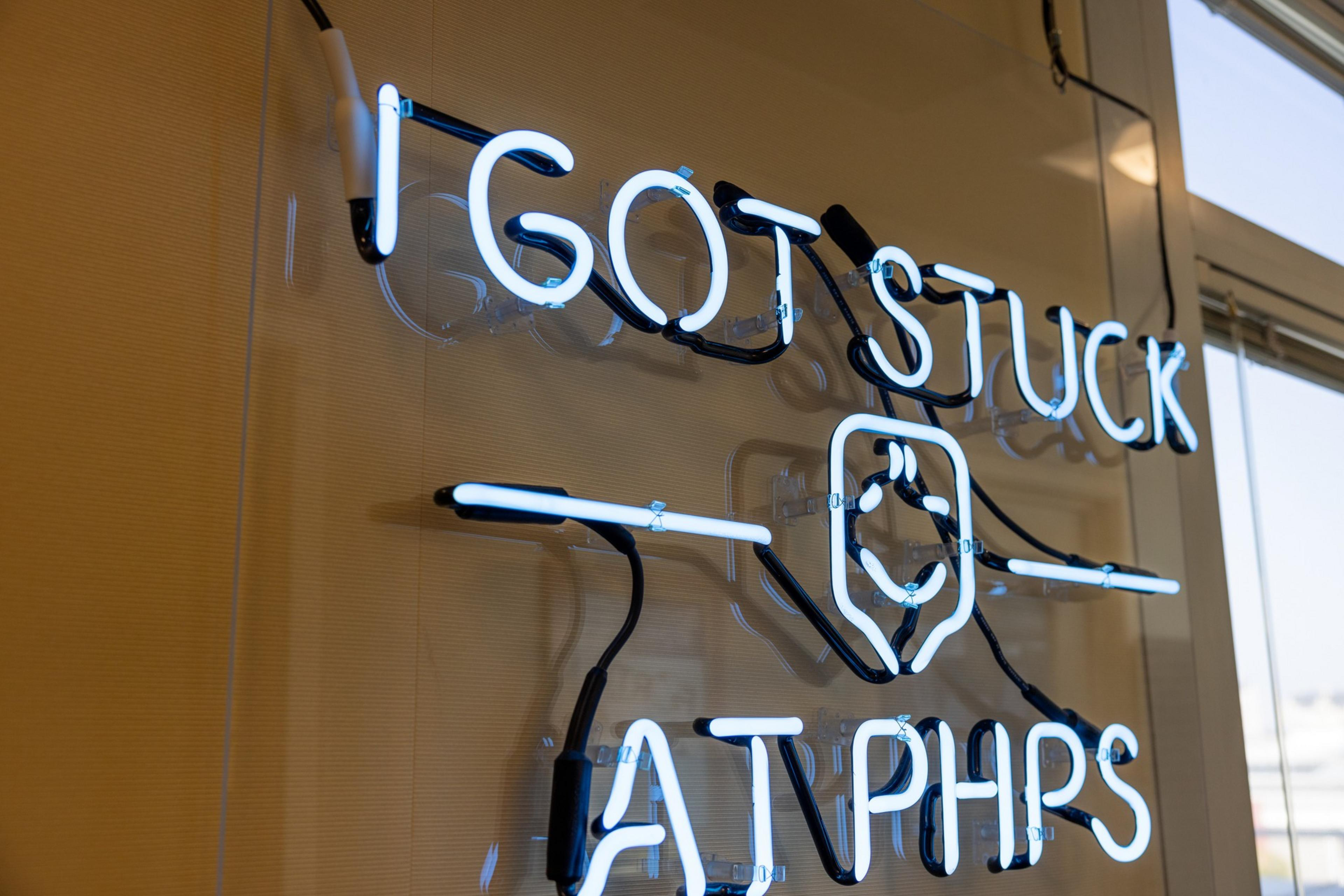 White neon sign on a wall reads "I GOT STUCK AT PHPS" with a simple outline of a smiling face between the words.