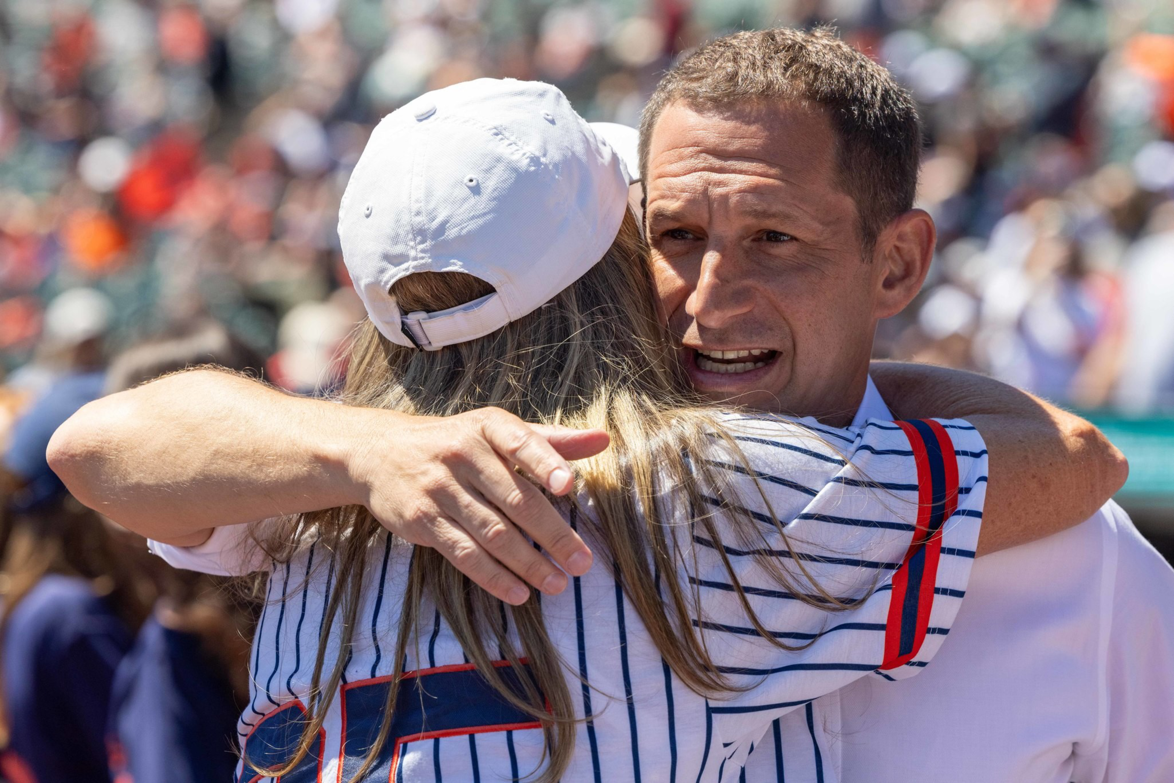 A man with short hair hugs a woman wearing a white cap and striped shirt, with a crowd visible blurred in the background.